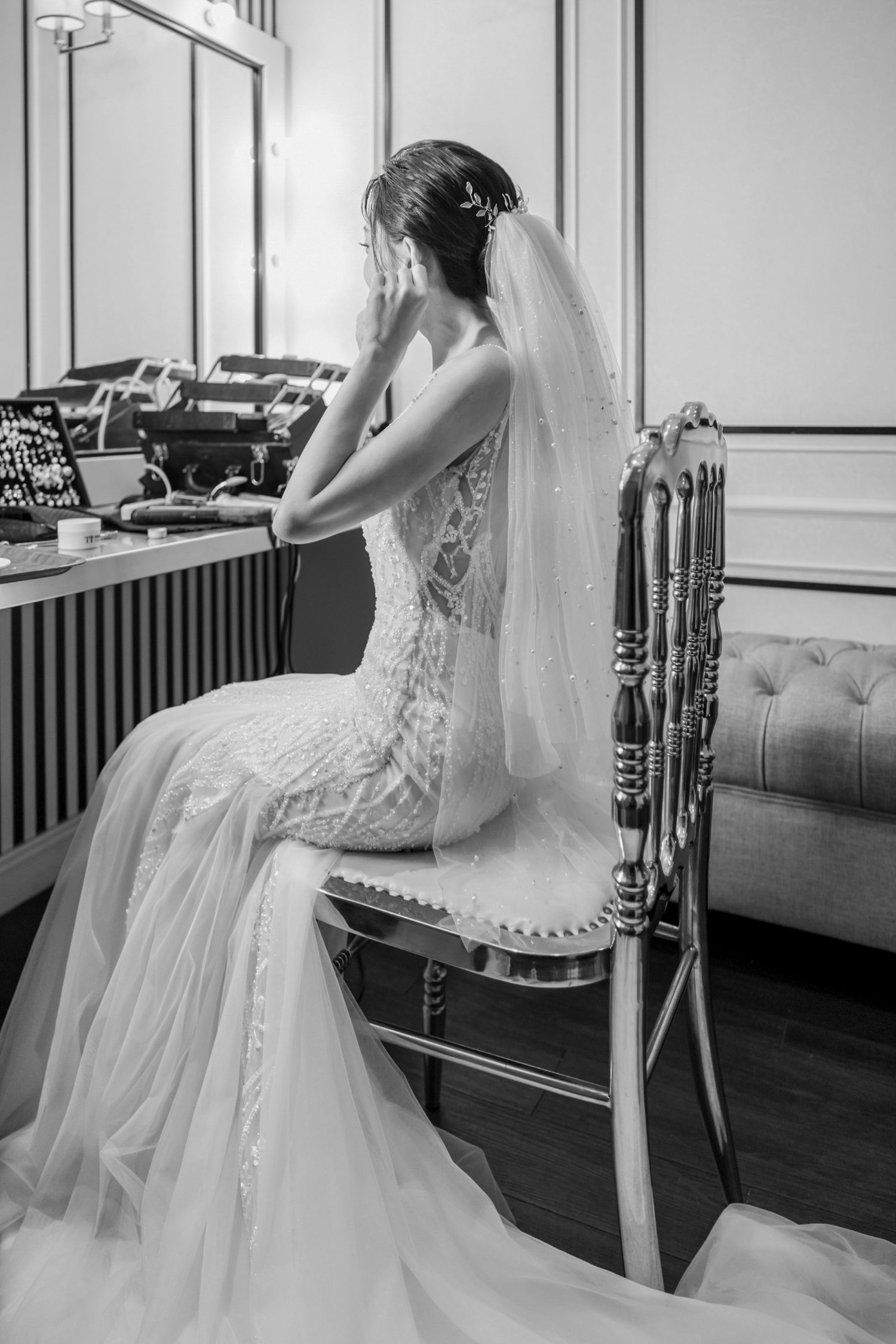 Black and white wedding photo showing bride in lace gown with flowing veil seated at vintage vanity table.