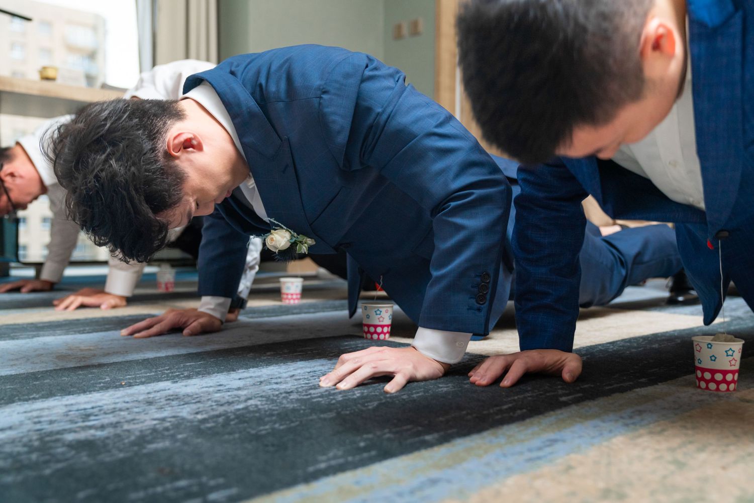 Two men in dark blue suits perform push-ups at what appears to be an office or workplace setting.