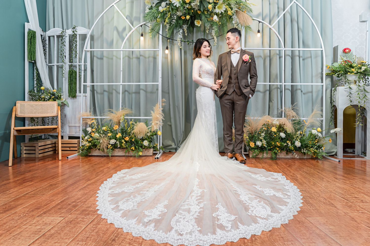 A wedding couple poses in front of an elegant white iron arch decorated with greenery on a hardwood floor.