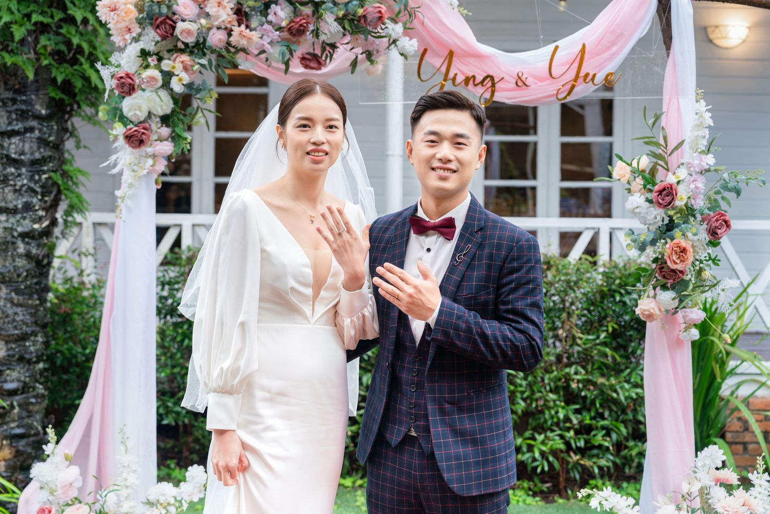 A joyful wedding moment captured under a floral arch decorated with pink and white roses at an outdoor ceremony setting.