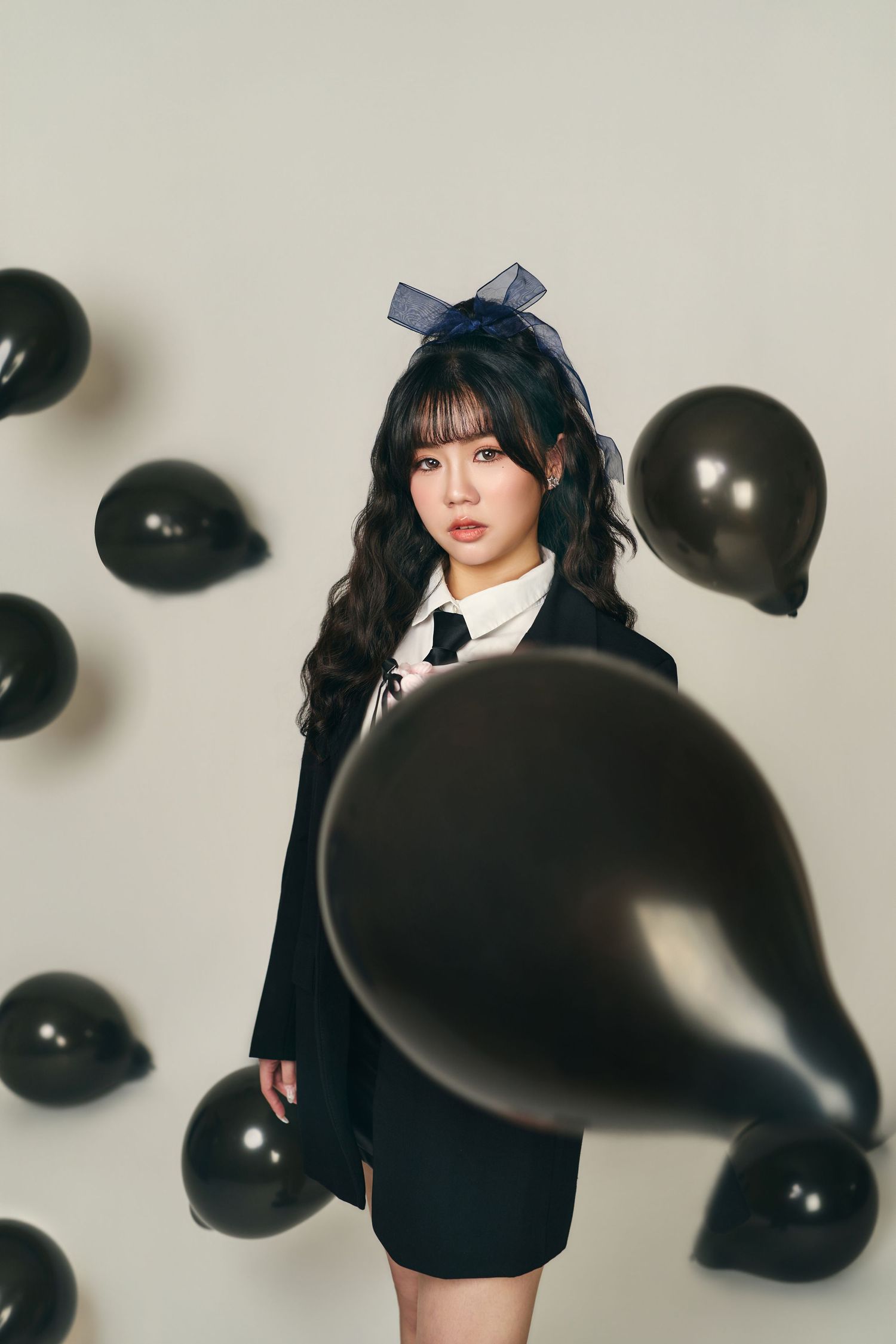 A person in a black outfit poses against a white backdrop with floating black balloons creating a dramatic aesthetic.