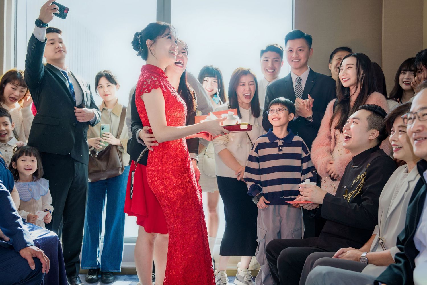A festive gathering shows a person in a red traditional Chinese dress performing a tea ceremony while guests watch and take photos.