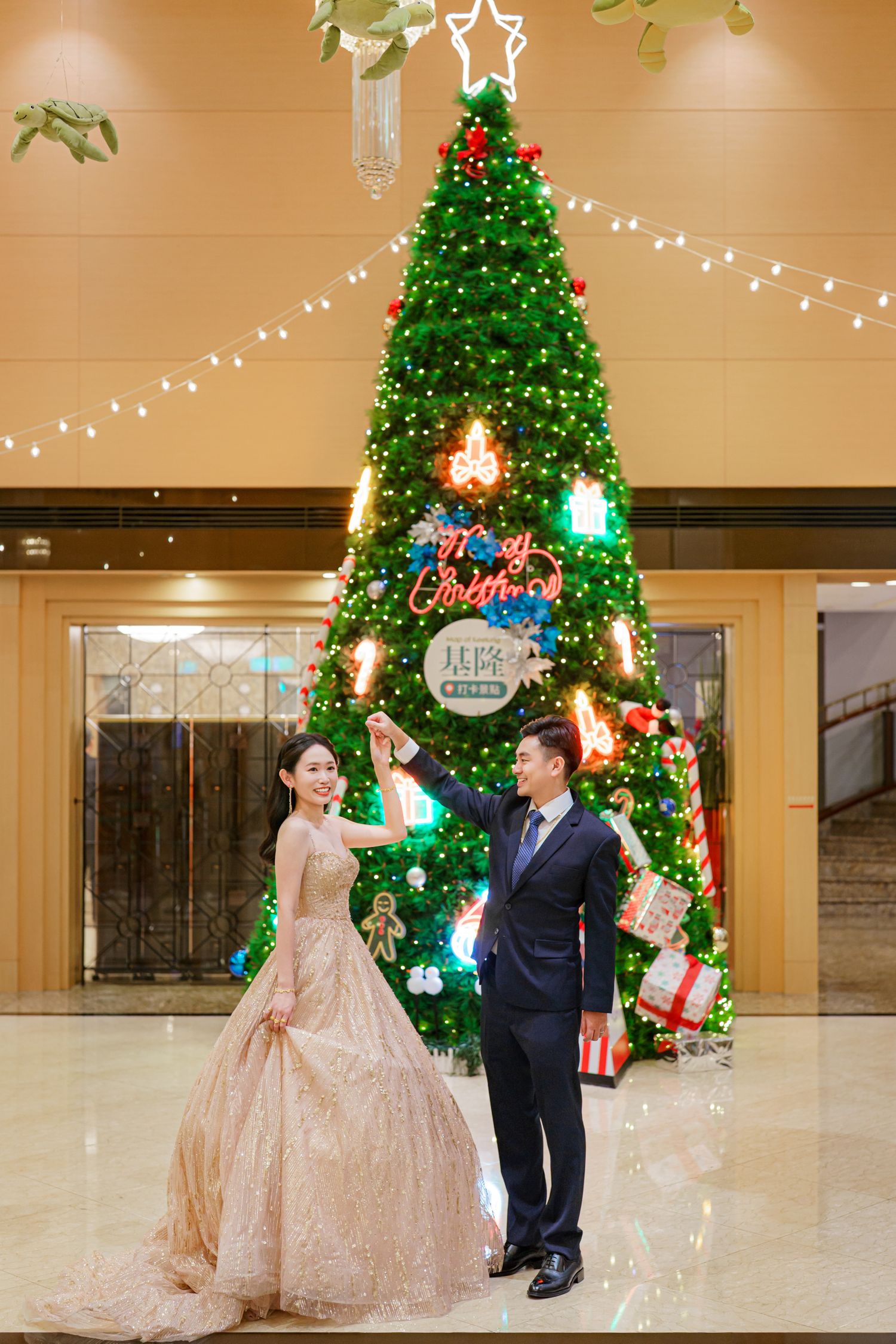 A couple in formal attire dances together in front of decorated Christmas trees in an elegant ballroom setting.