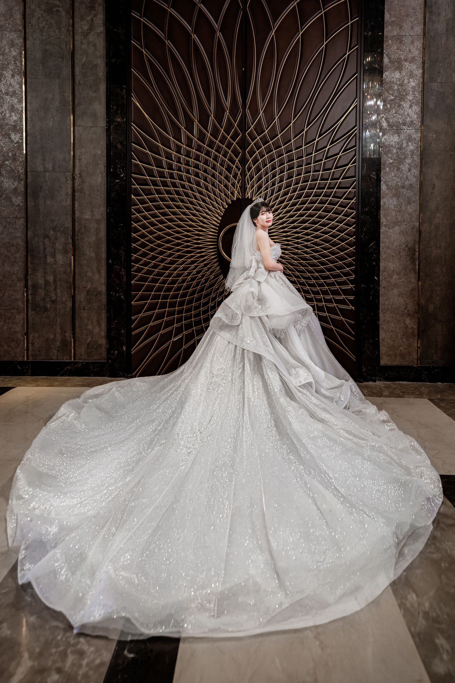 Bride in a stunning white ballgown with long glittering train and veil, posed before an ornate gold Art Deco wall panel.
