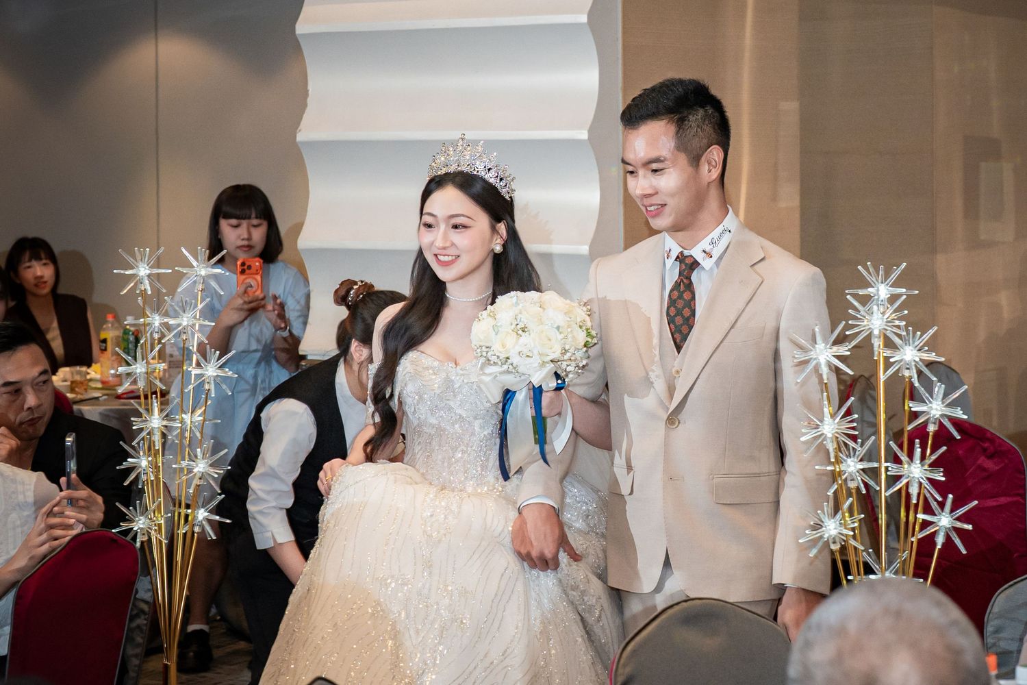 Bride in white lace gown and tiara walks with groom in beige suit at elegant wedding ceremony reception.