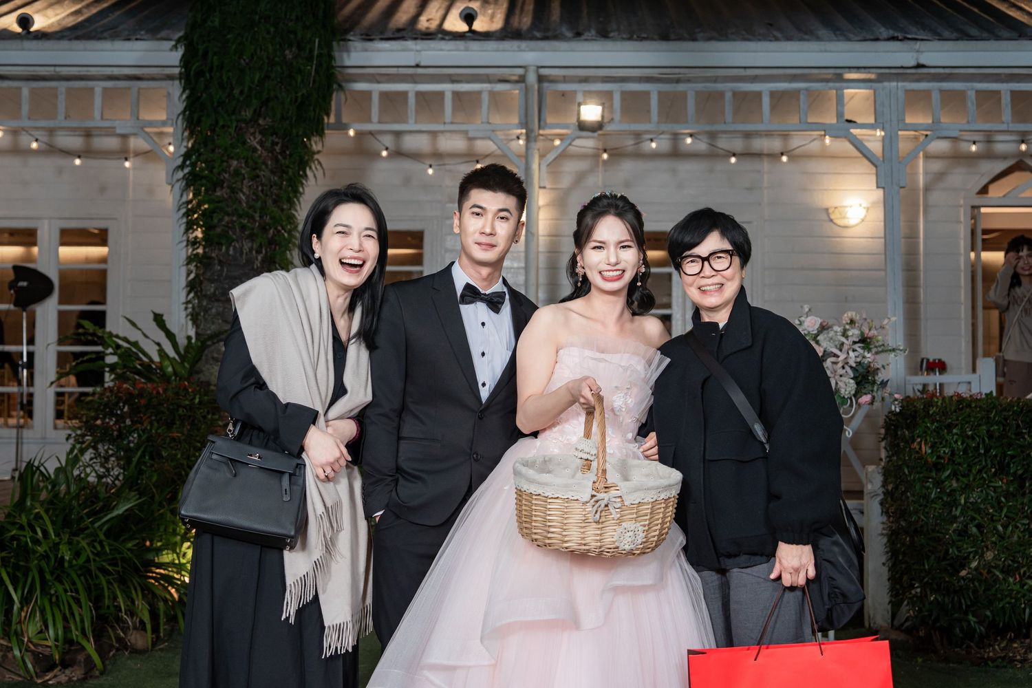Wedding celebration group photo in front of a white building with decorative lighting and landscaping.