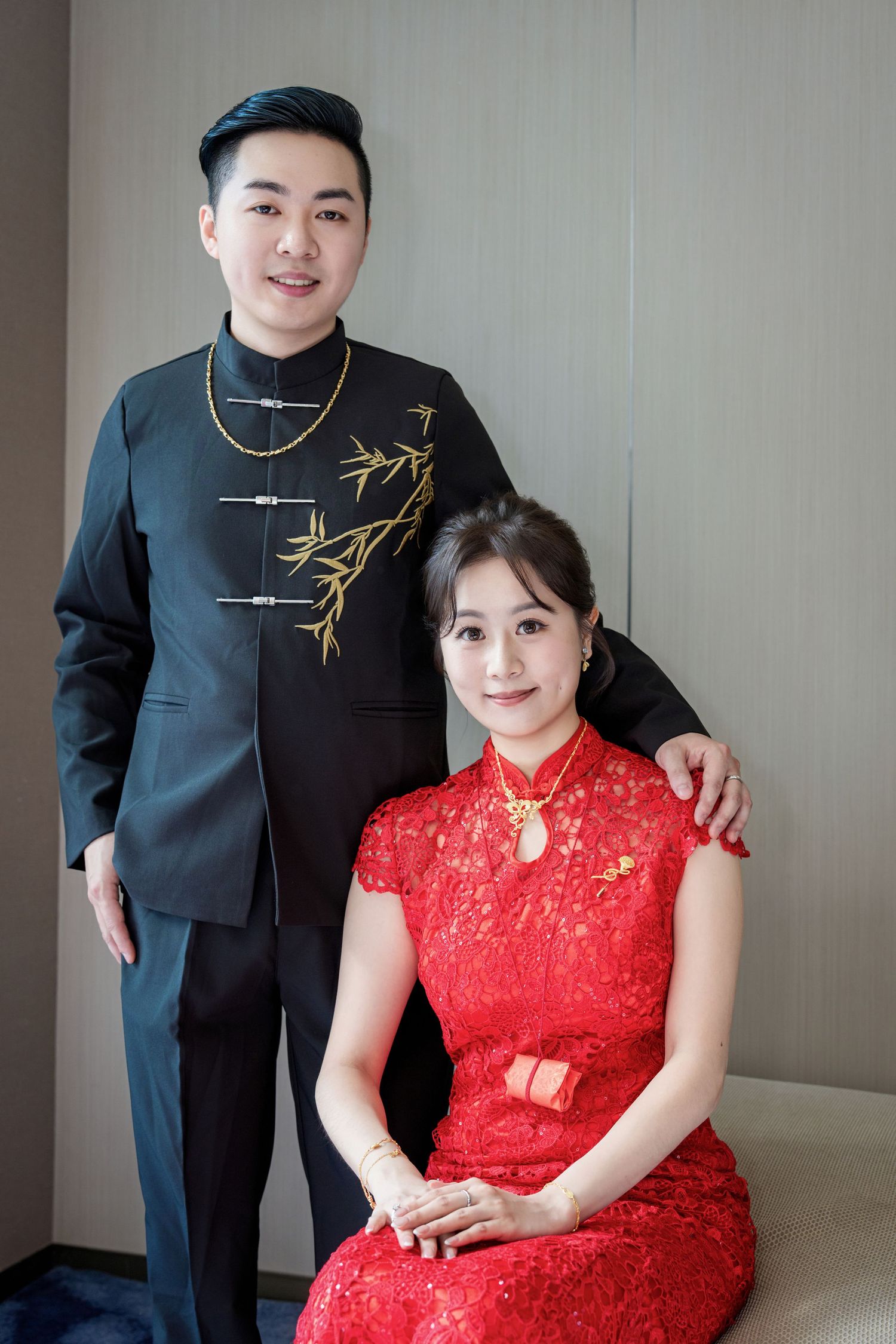 A couple in traditional Asian formal wear with a black mandarin collar suit and red lace qipao dress pose together.