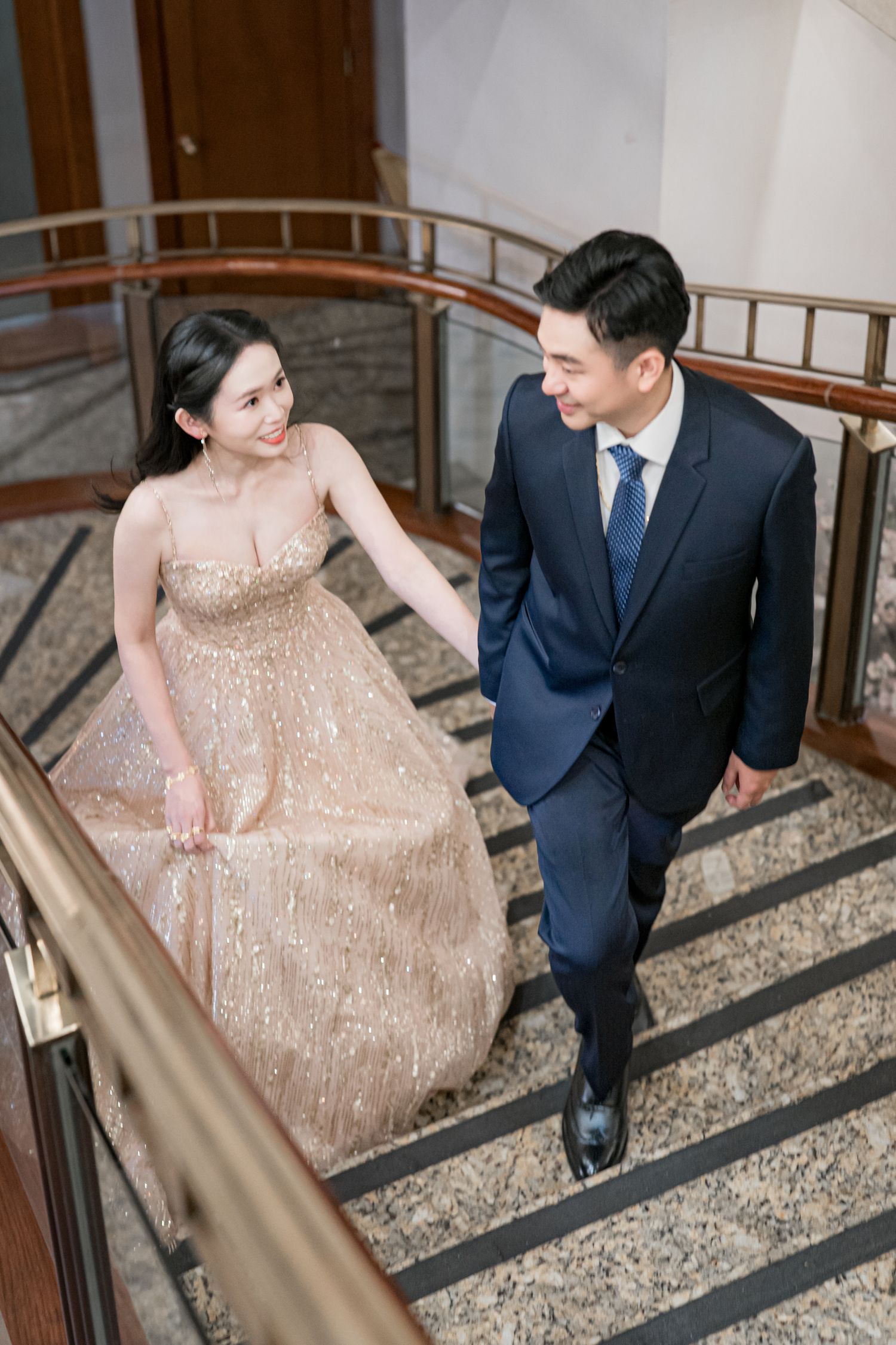 A couple descends an elegant staircase together, with the bride wearing a sparkling champagne-colored gown.