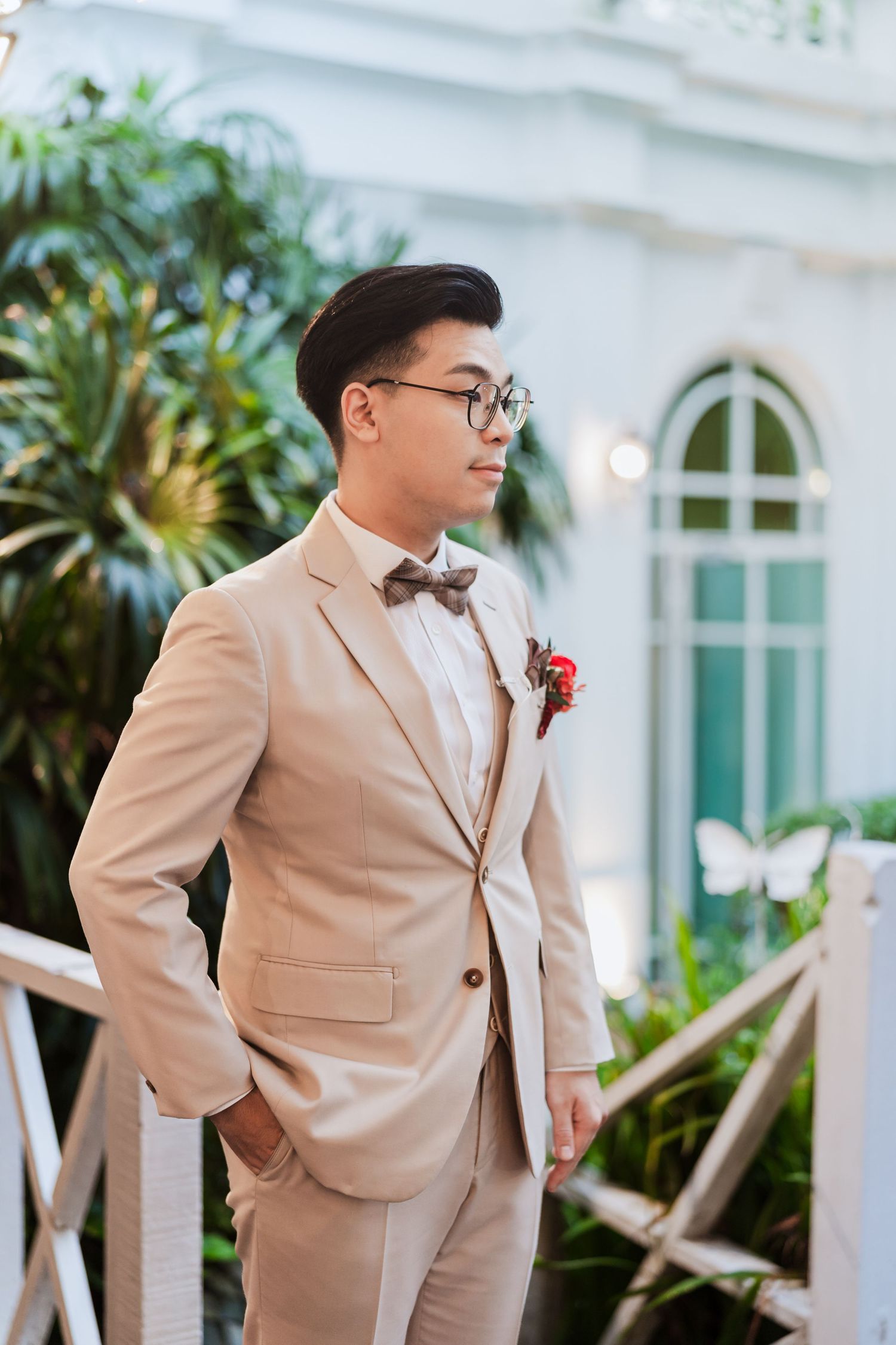 A groom in a beige suit and bow tie stands elegantly against a tropical backdrop with arched windows.