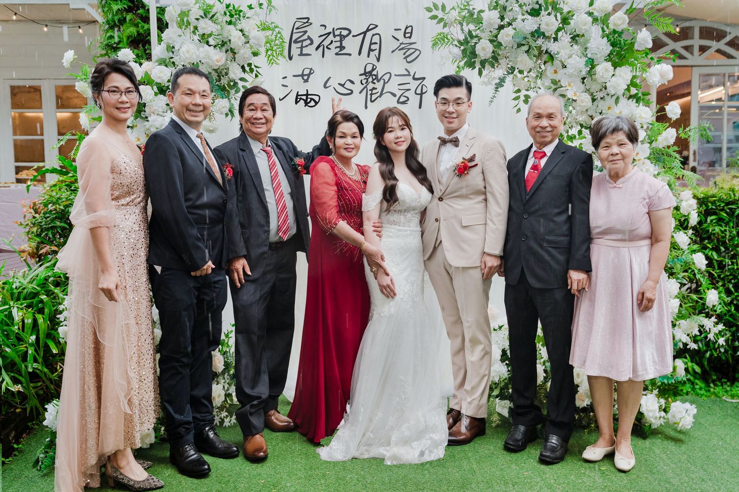 Wedding party poses together under elegant white floral arch against greenery backdrop at outdoor garden ceremony.