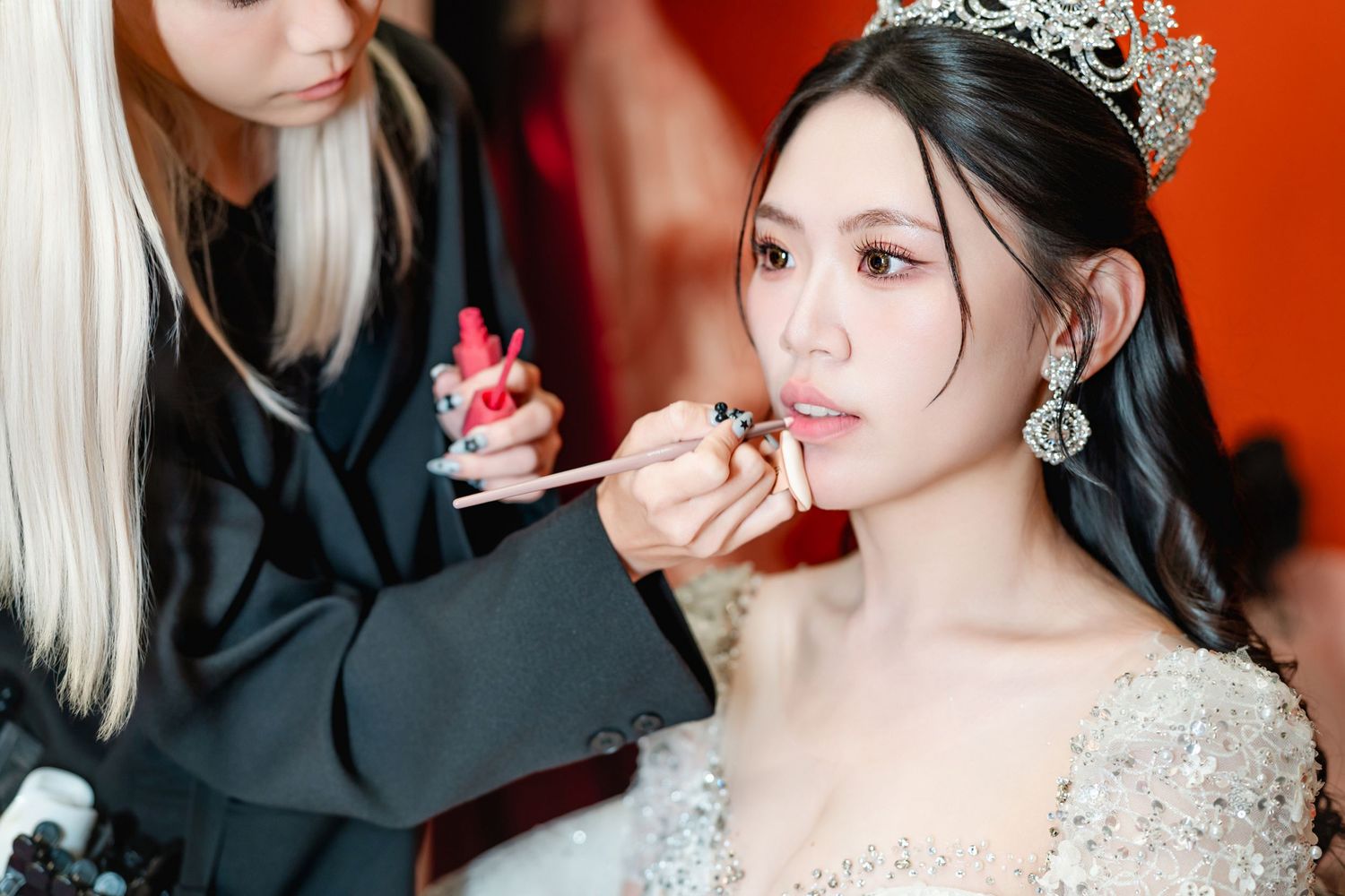 A bride in a sparkling gown and crystal crown has makeup applied by a stylist against a red background.