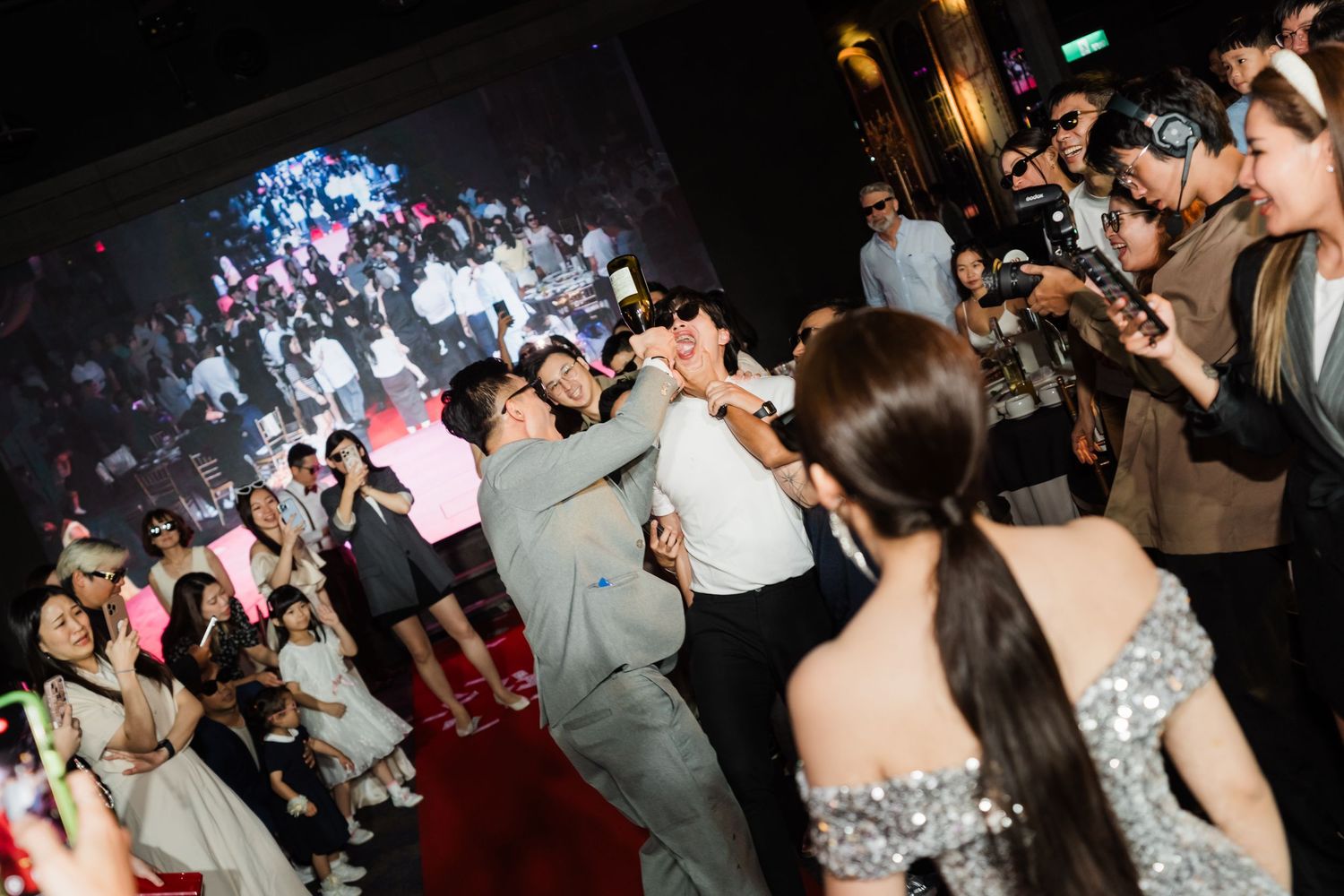 A crowded dance floor at a nighttime party or celebration with people dancing under colorful lights and capturing photos.