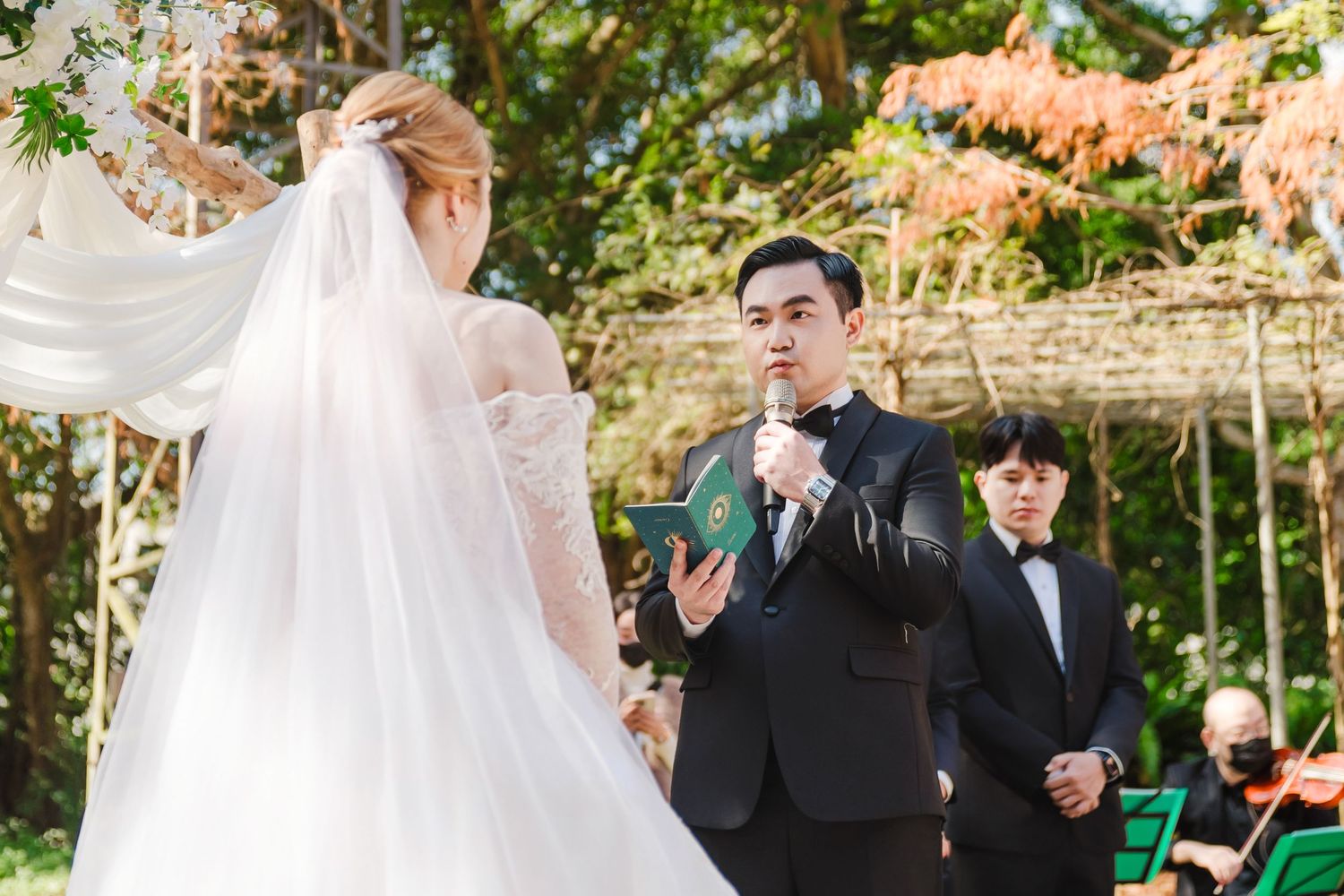 An outdoor garden wedding ceremony with a flowing white bridal veil and guests gathered under floral arrangements.