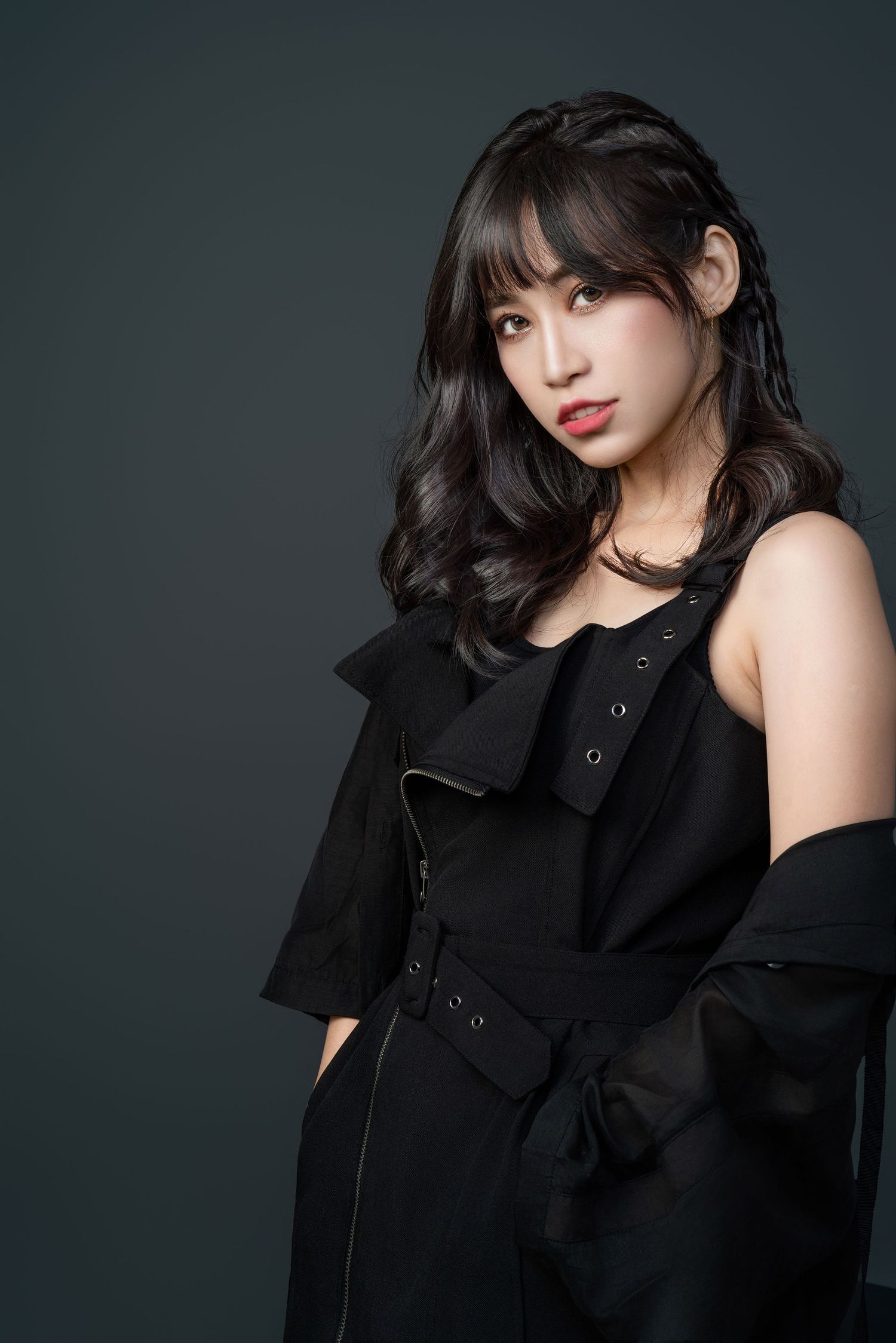 Model in elegant black layered outfit with dark wavy hair poses against dark background in studio portrait.