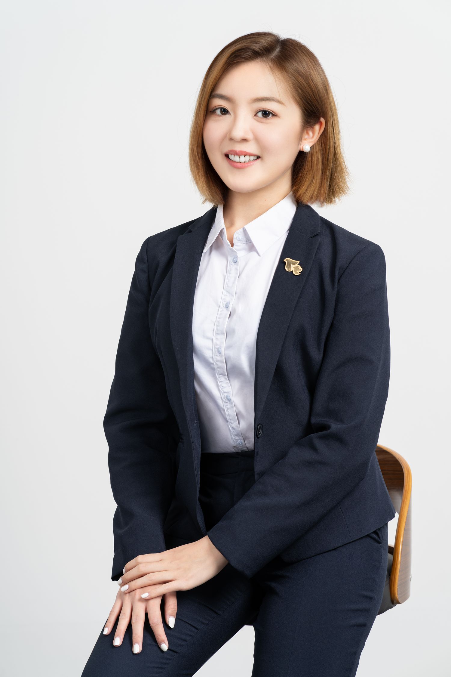 Professional in navy business suit seated on wooden chair against white background with small decorative pin on lapel.