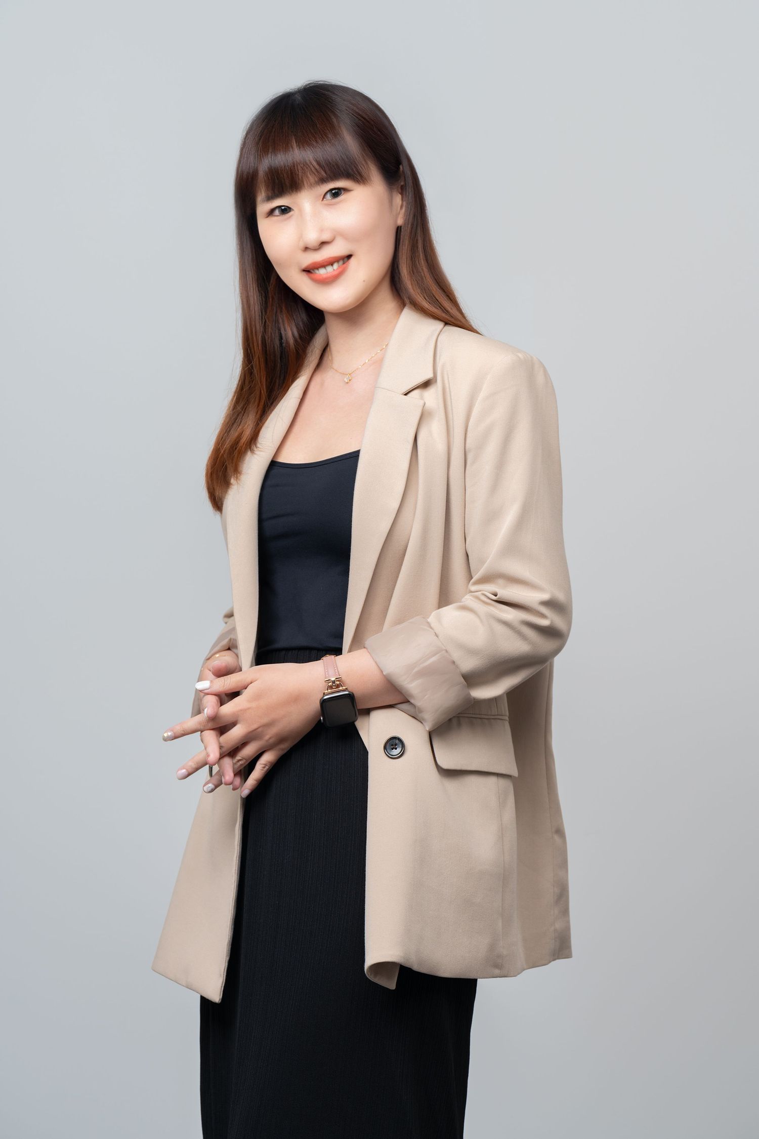 Professional woman in business attire wearing a beige blazer over a black dress stands against gray background.