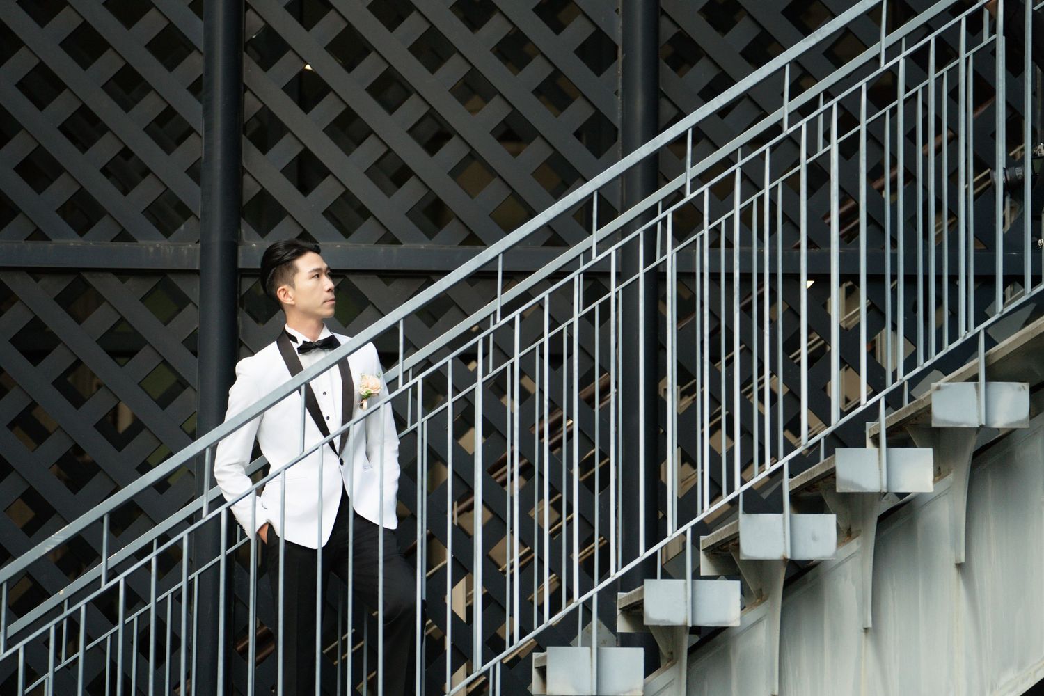 A person in a white blazer leans against a modern metal staircase railing against a dark latticed wall.