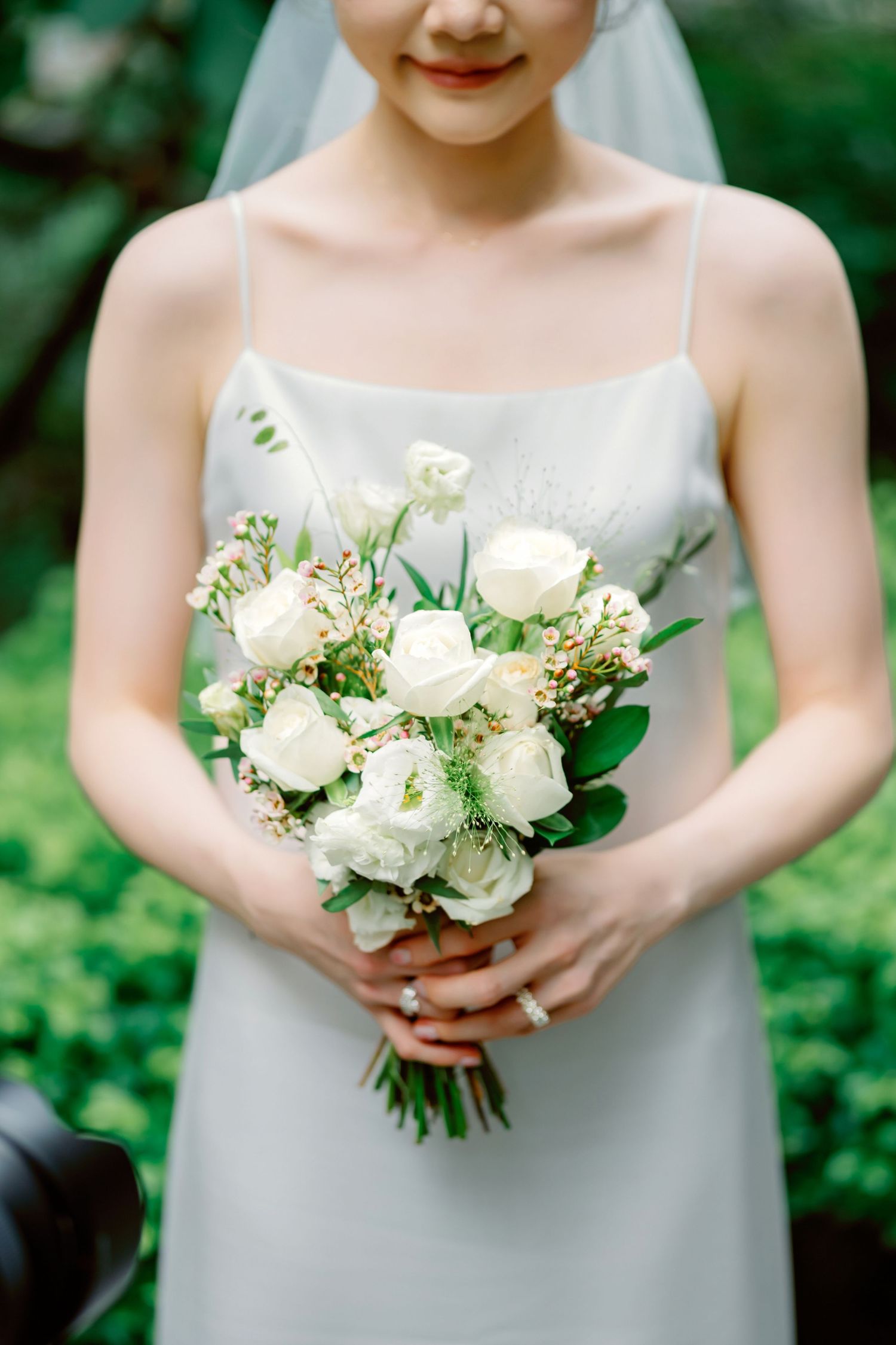 Elegant white rose and baby's breath wedding bouquet held by bride in simple white spaghetti strap dress against green garden.