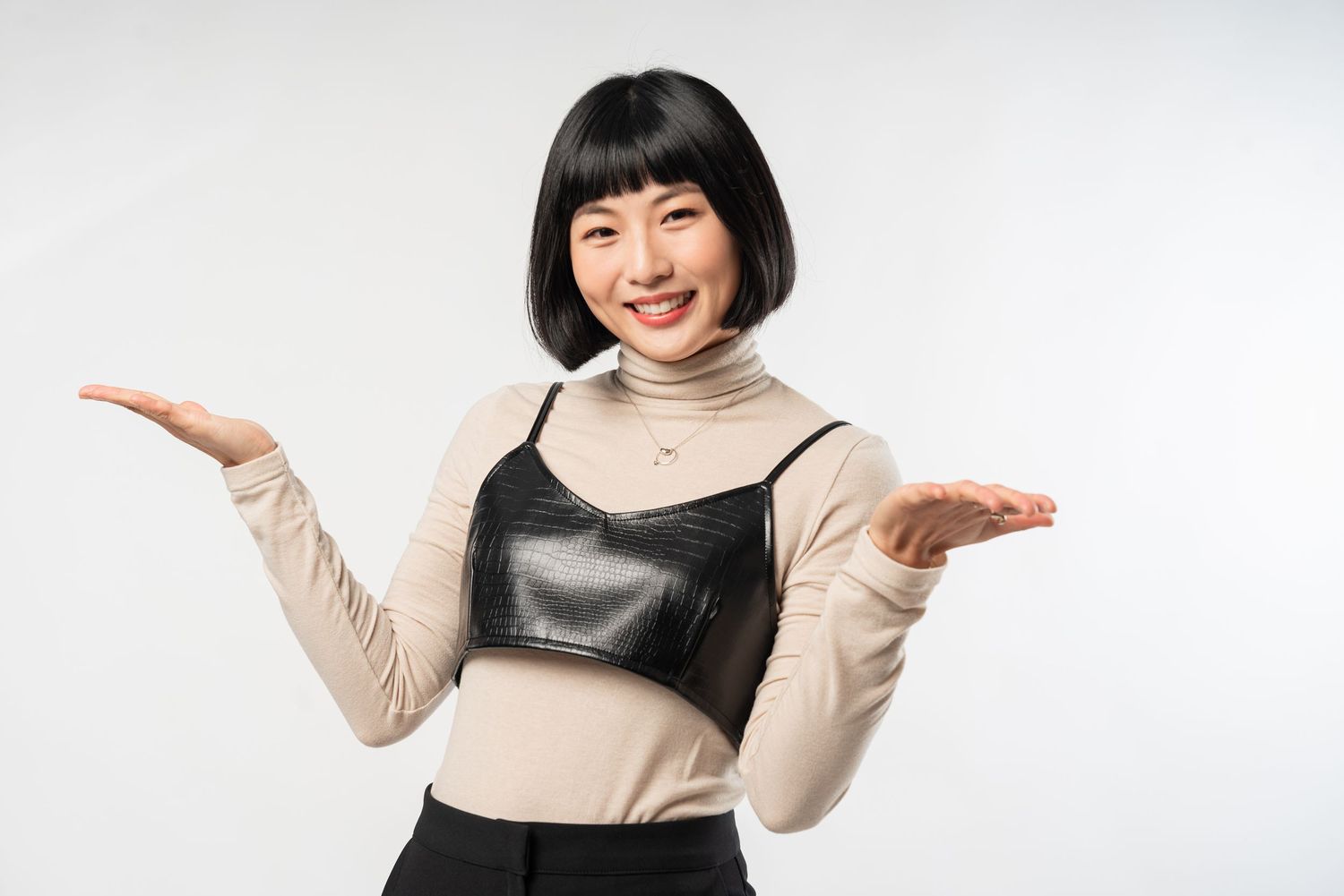 Smiling person in beige turtleneck sweater and black dress making expressive hand gestures against white background.