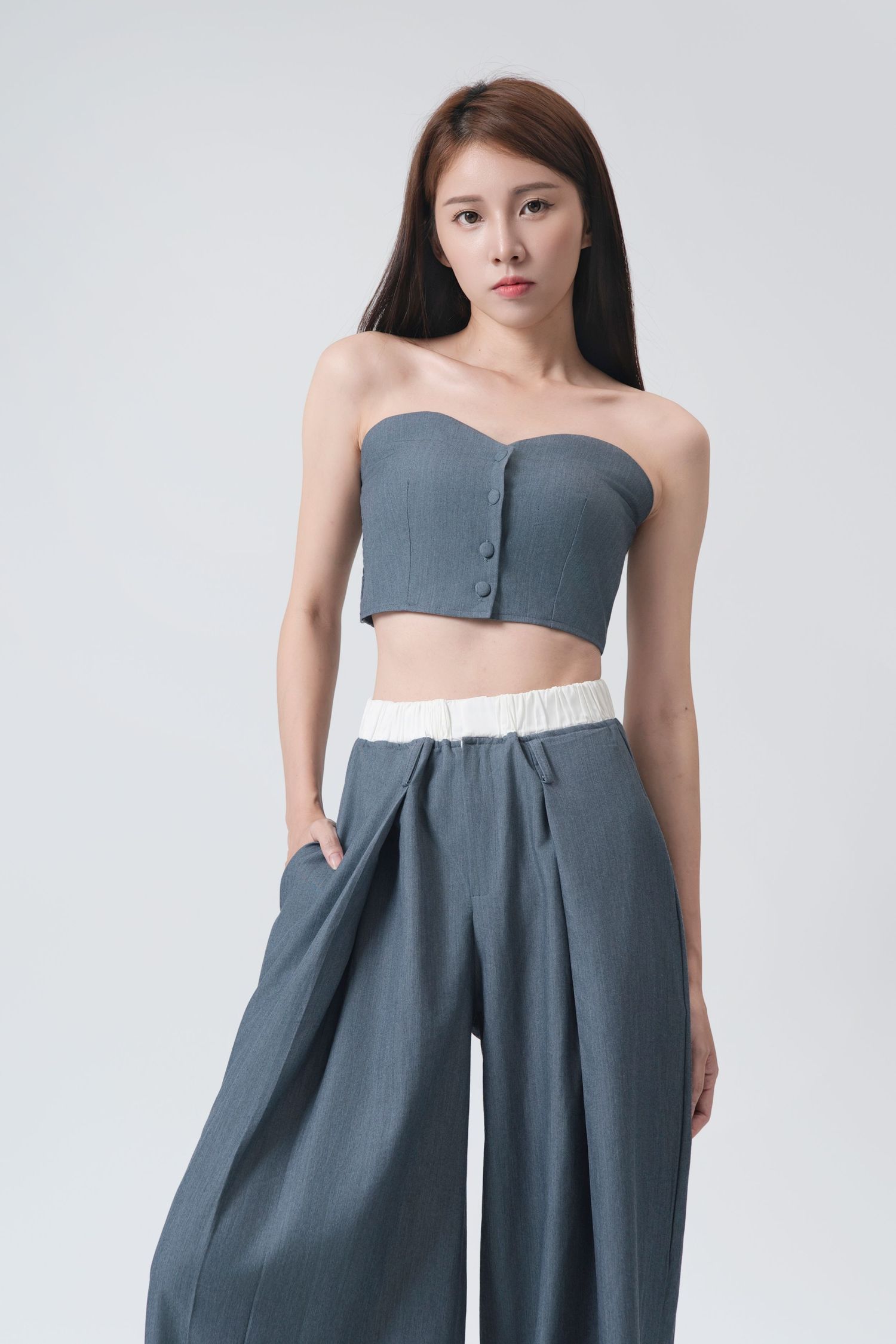 Model wearing a grey strapless crop top and high-waisted wide-leg pants in a minimalist studio setting.
