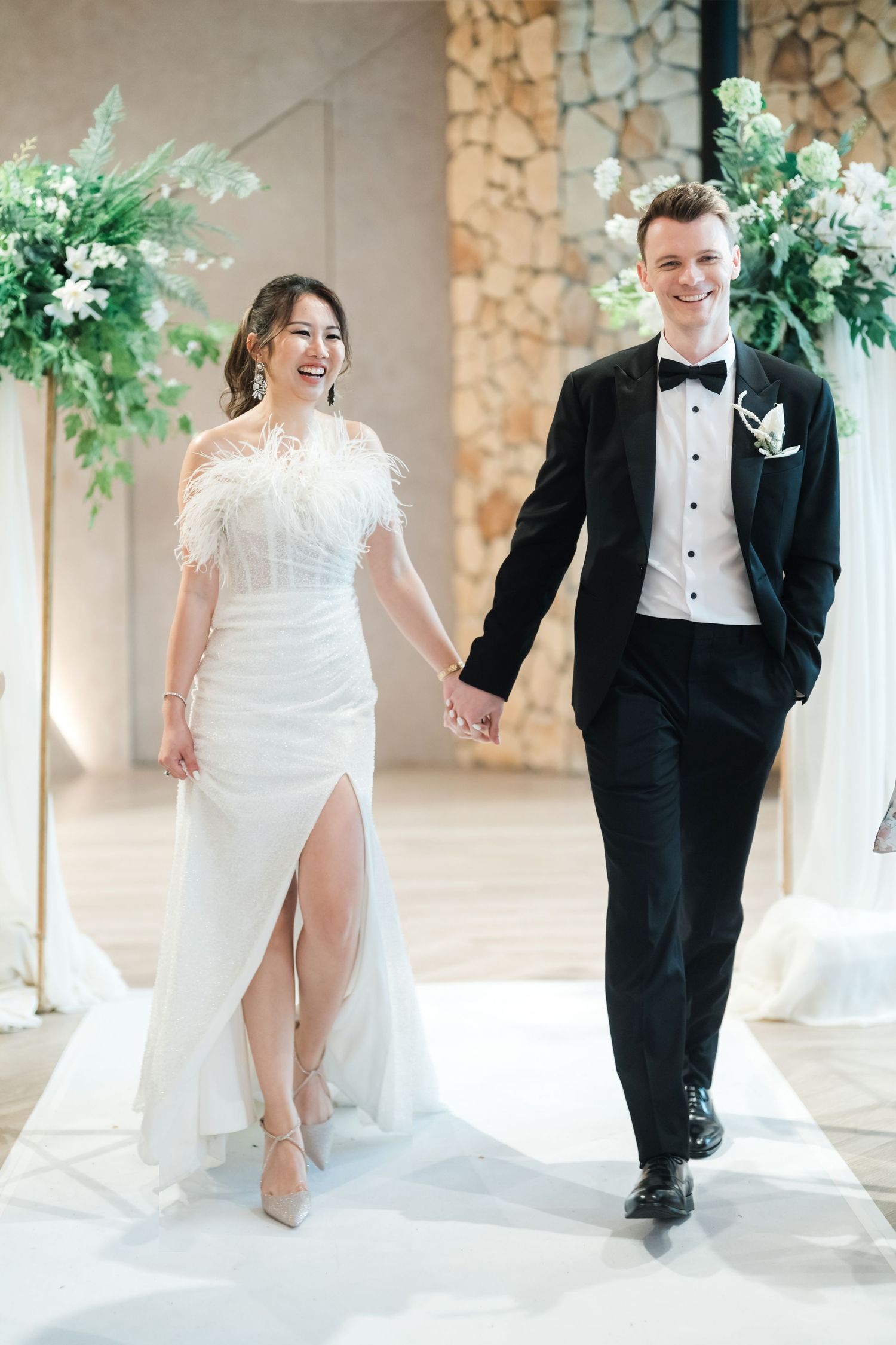 A bride and groom run and laugh together in a bright hallway with stone pillars and floral decorations.