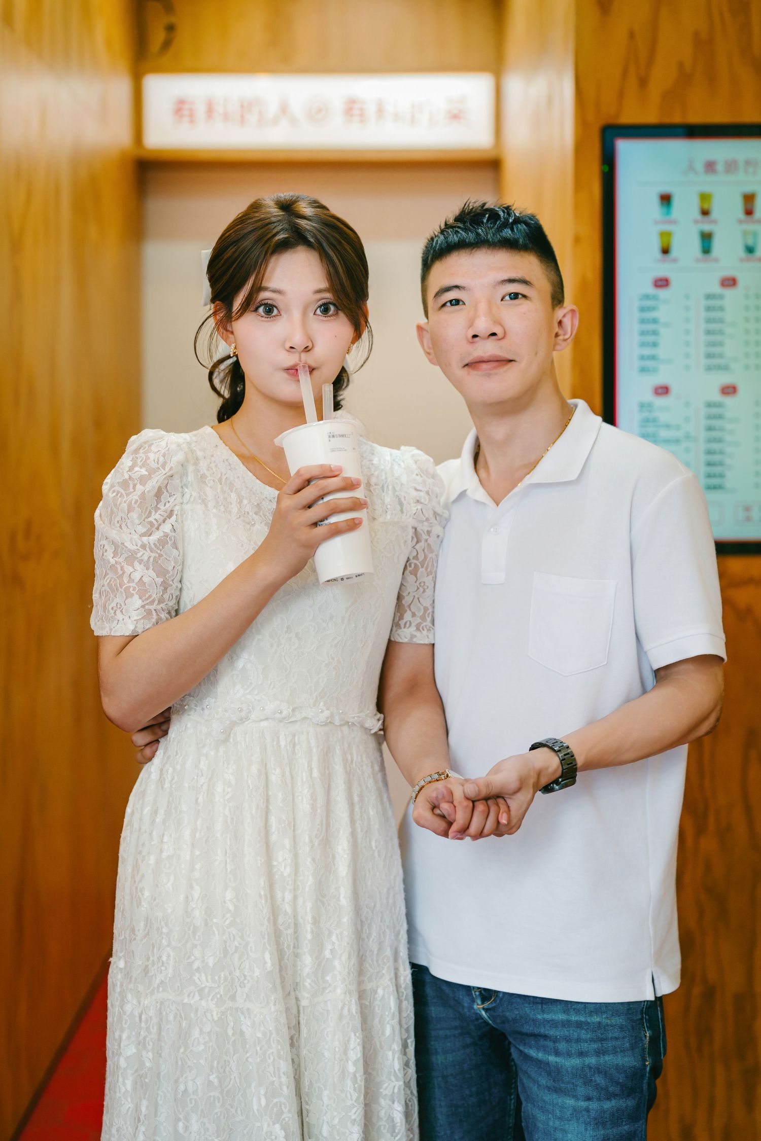 Two people in casual attire stand together drinking beverages against a wooden wall background.