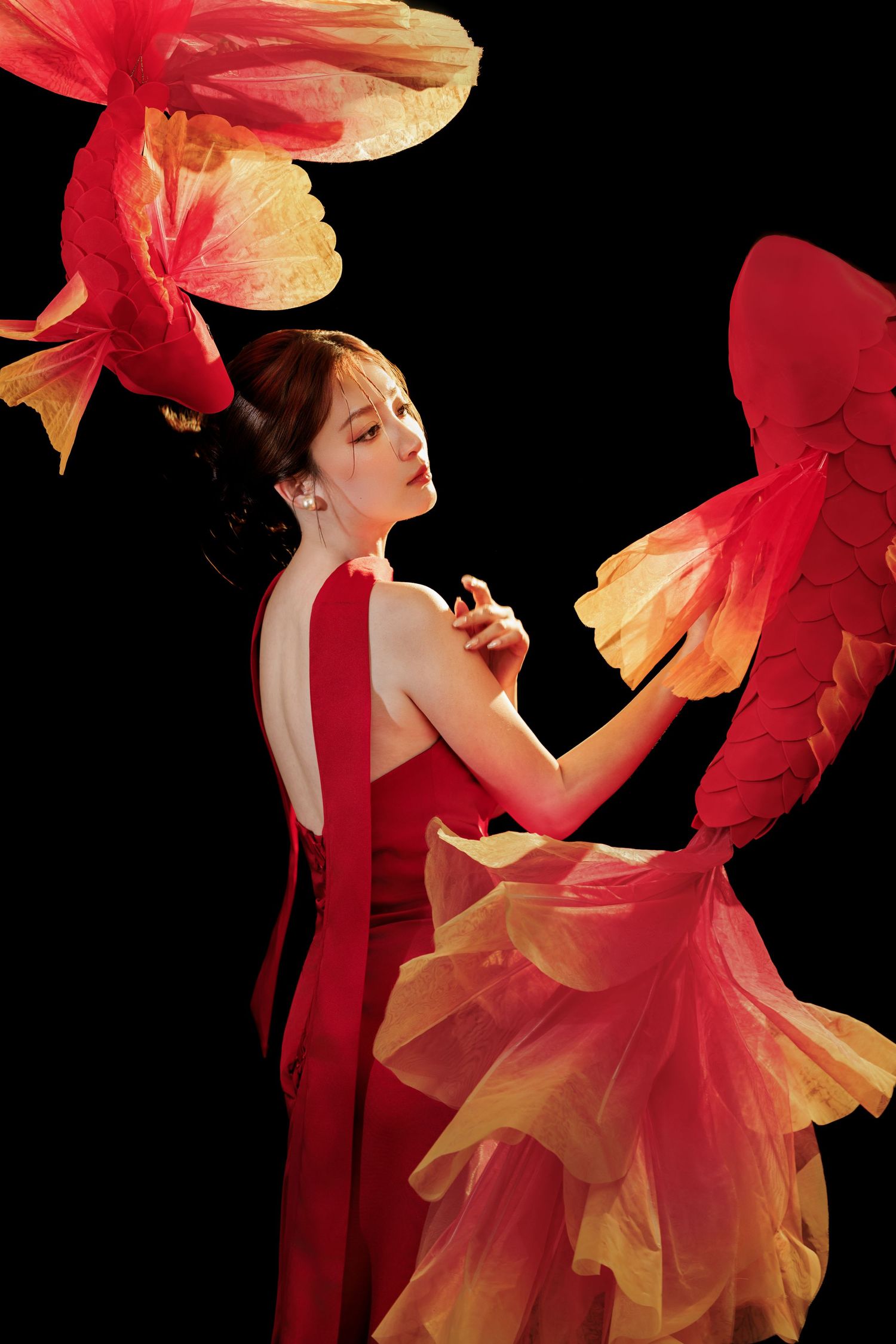 A woman in a red backless gown poses gracefully with vibrant red and gold koi fish decorations on black background.