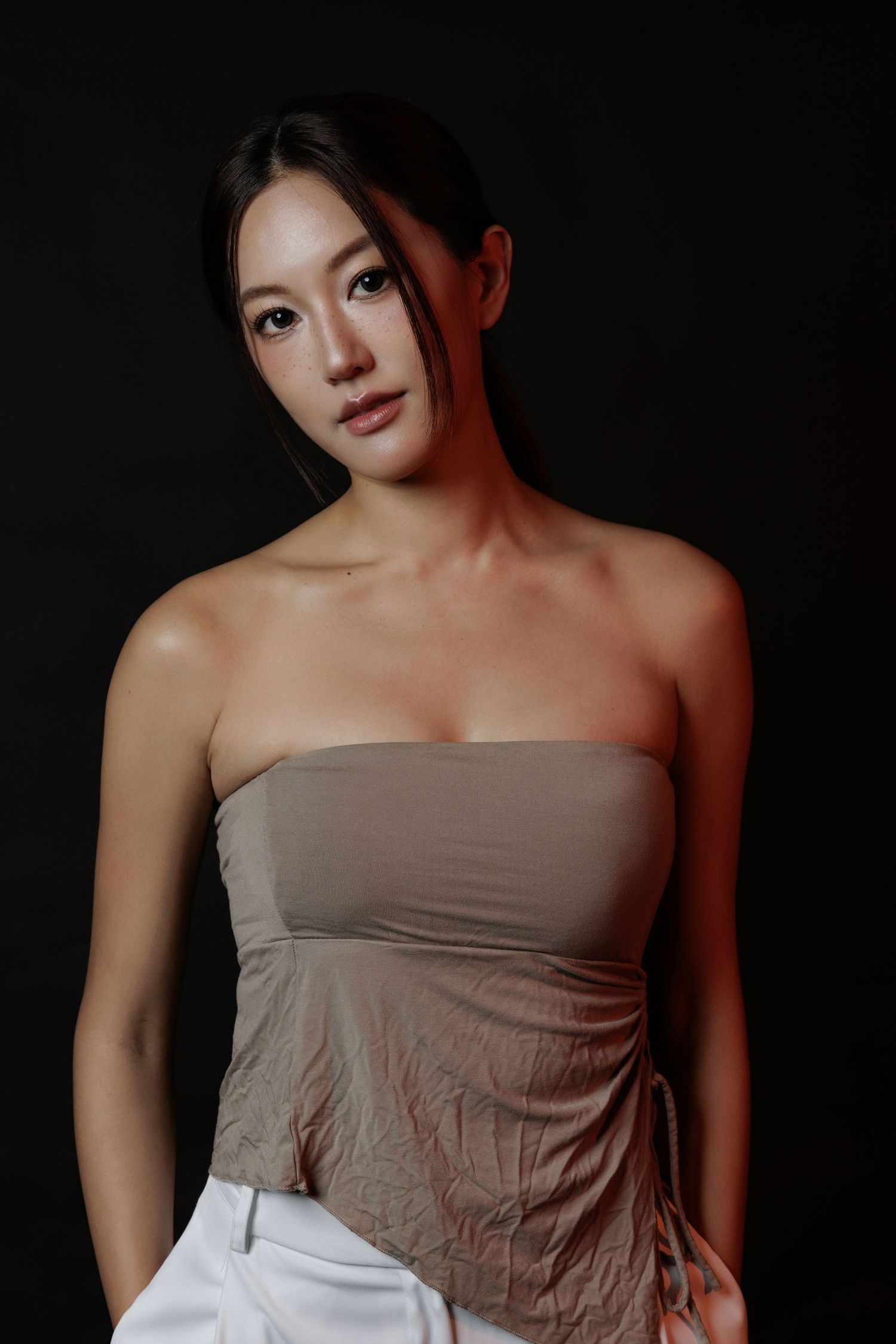 A young Asian woman poses in a taupe strapless top and white pants against a dark background with dramatic lighting.