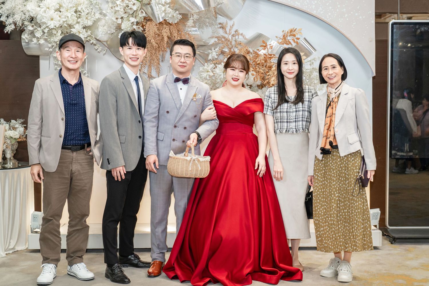 A group of wedding guests pose together in formal attire including a red dress and gray suits at a celebration event.