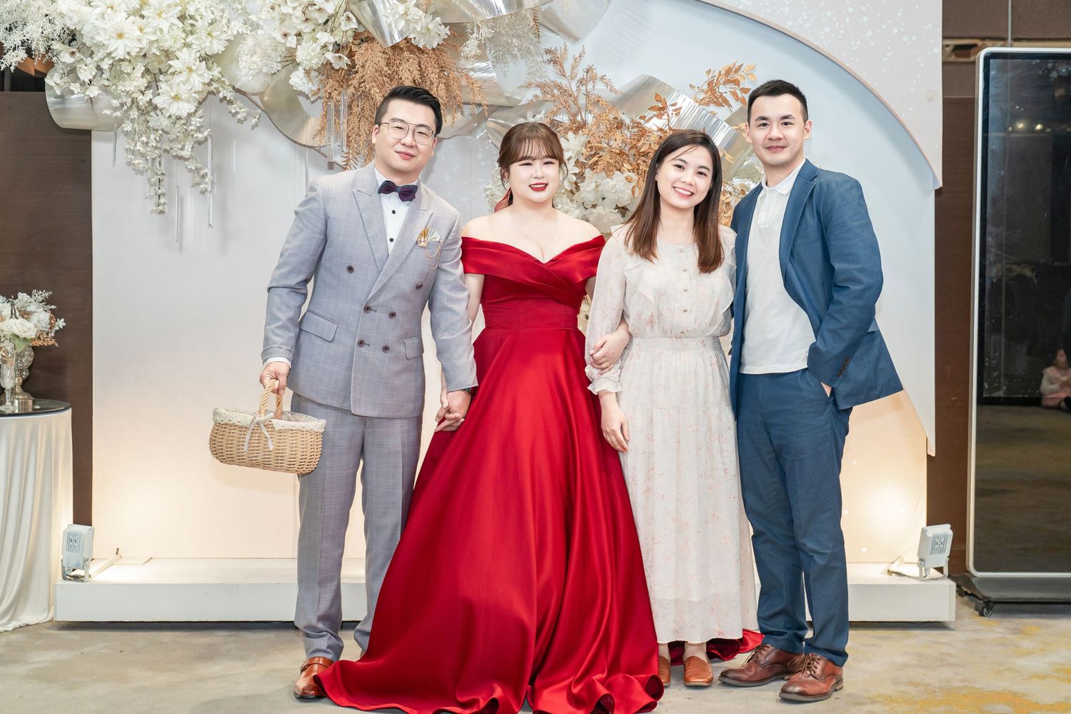 Wedding party members in formal attire pose together in an elegant white venue with floral decorations.