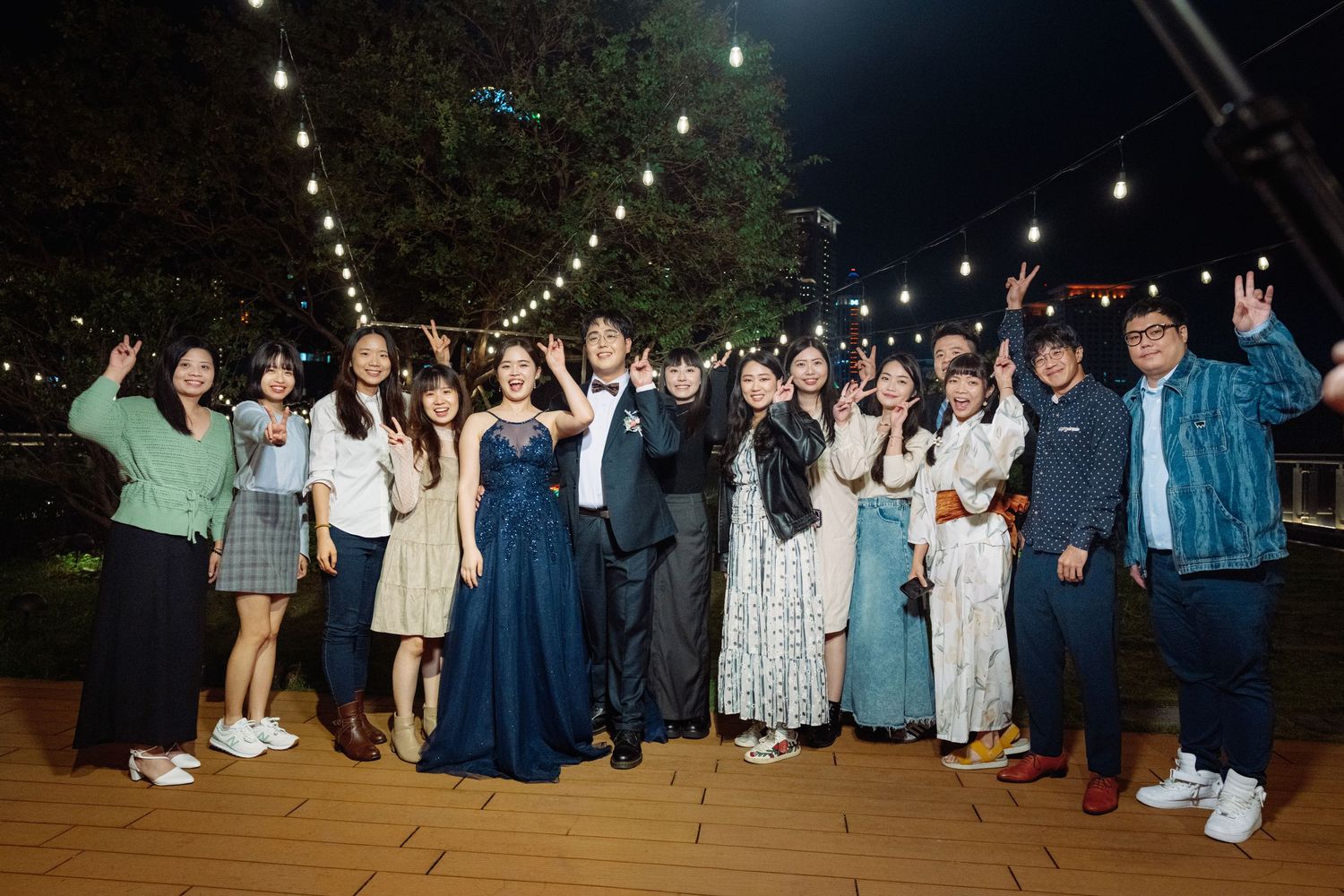 Wedding guests dance together on a wooden deck under string lights during an outdoor evening reception.