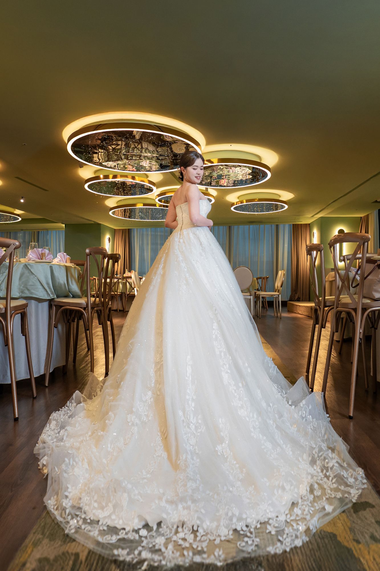 Elegant white wedding dress with lace details and long train displayed in a luxurious venue with modern lighting fixtures.