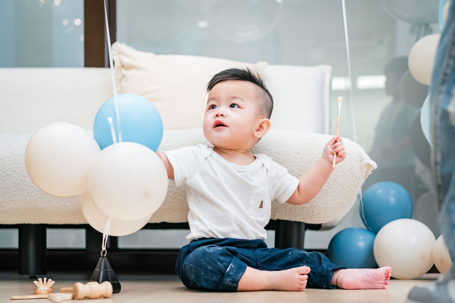 Baby in white shirt sits on wooden floor surrounded by blue and white balloons at first birthday party celebration.