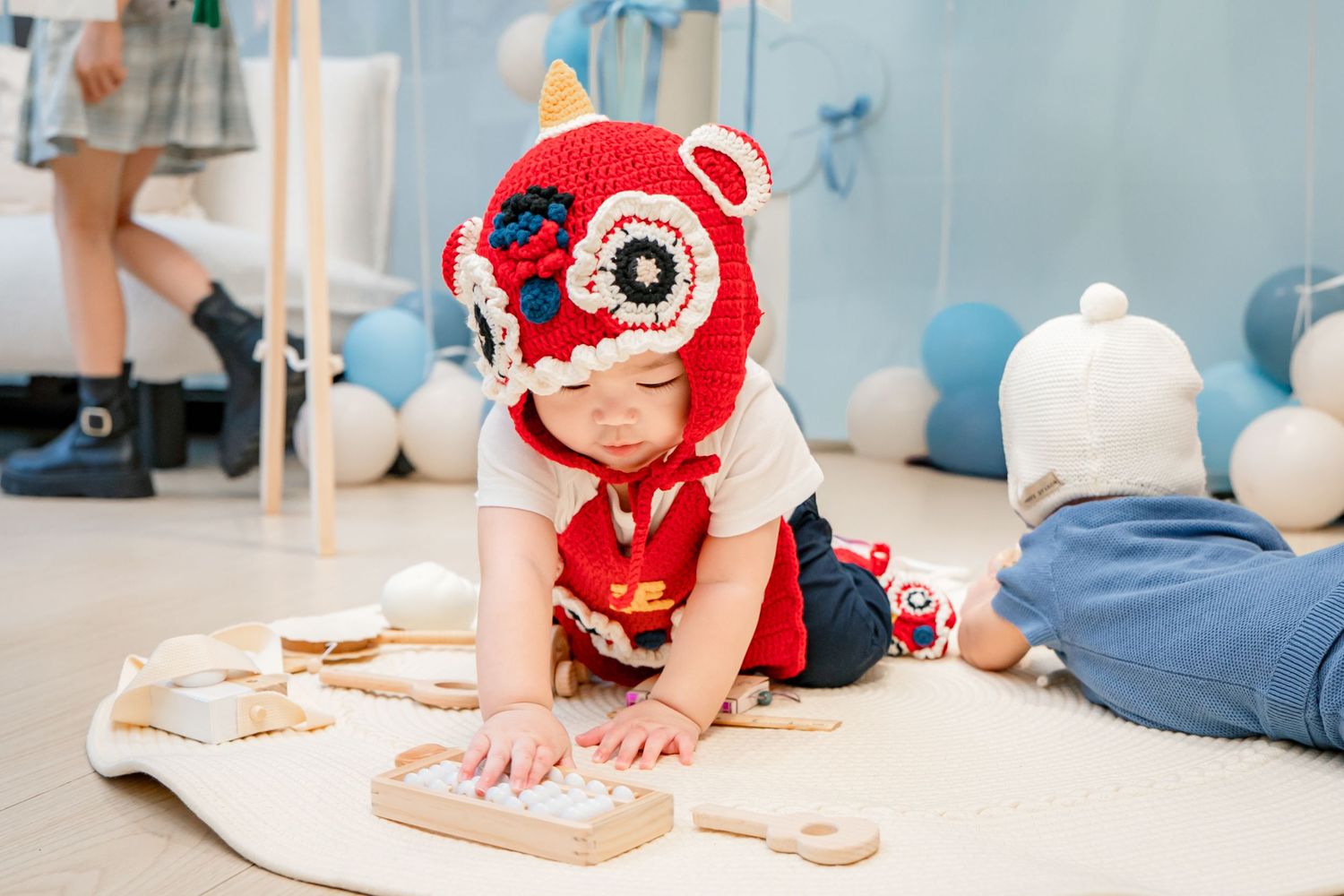 Baby wearing red and white knitted hat and white shirt crawls on floor next to cookies and baking supplies.