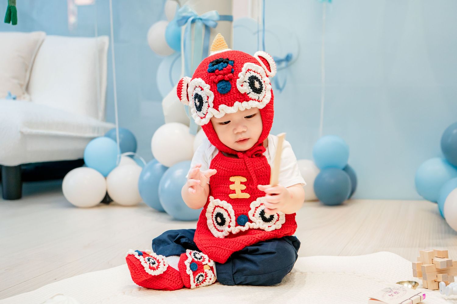 Baby wearing red Chinese lion dance hat sits on floor with blue and white balloon decorations in background.