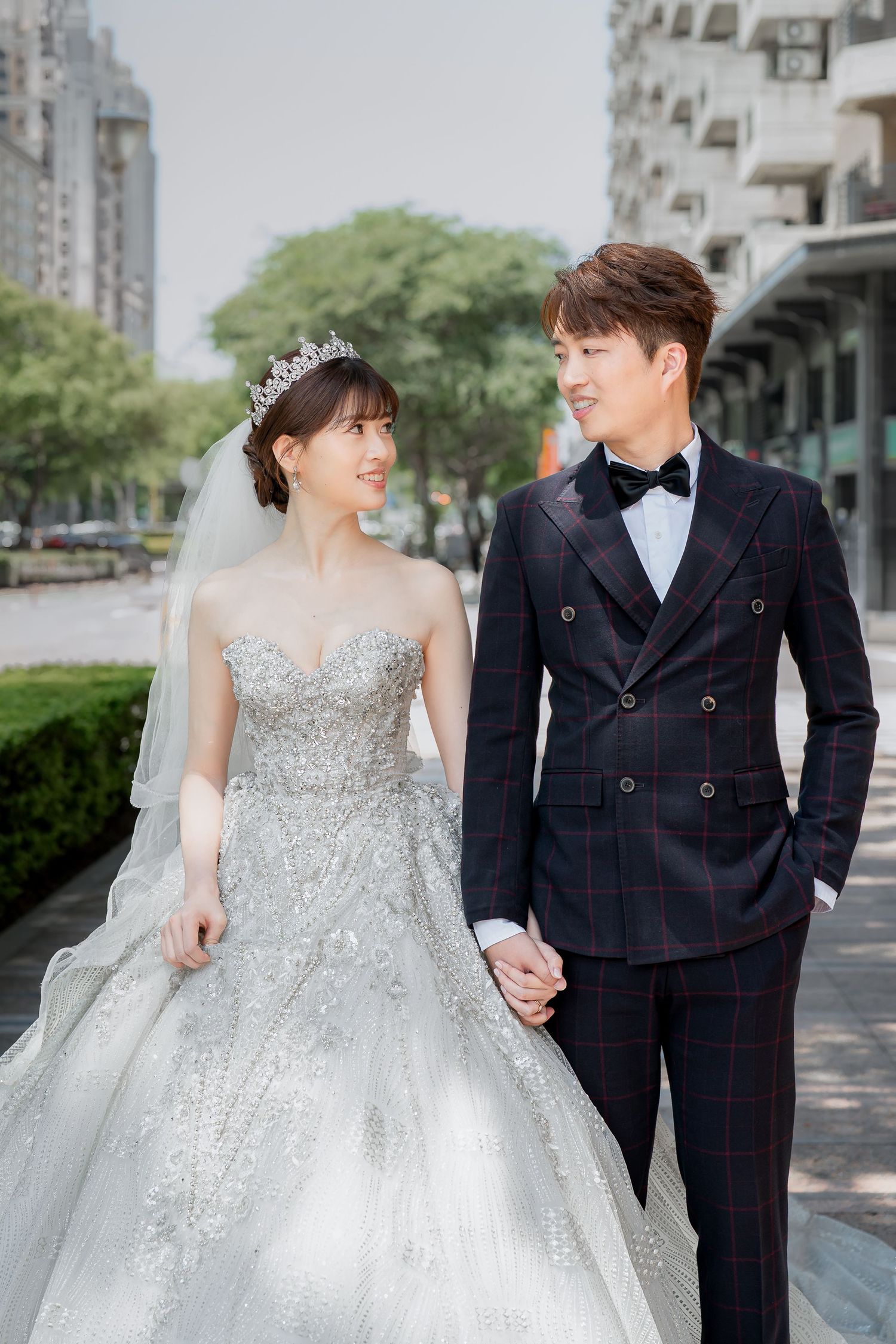 A couple in wedding attire holding hands outdoors, bride in beaded gown with tiara, groom in plaid suit with bow tie.