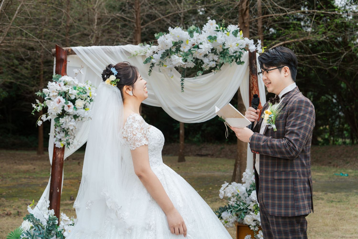 A couple in wedding attire stands outdoors near rustic wooden decor and floral arrangements during their ceremony.