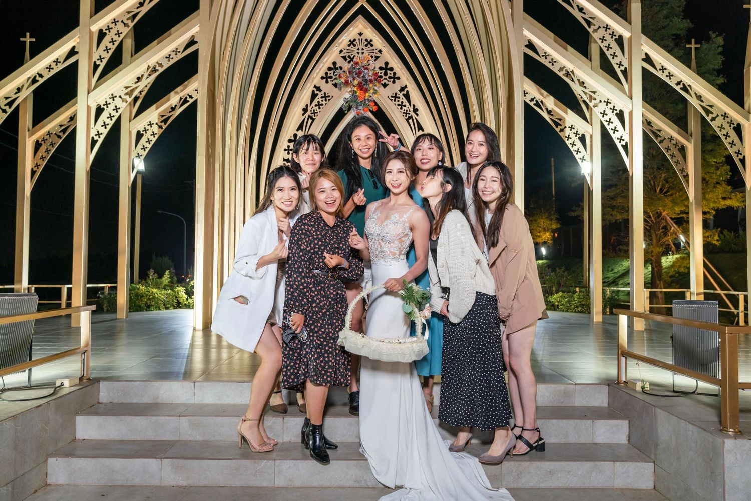 A wedding party poses together on the steps of a Gothic-style outdoor chapel with illuminated arches at night.