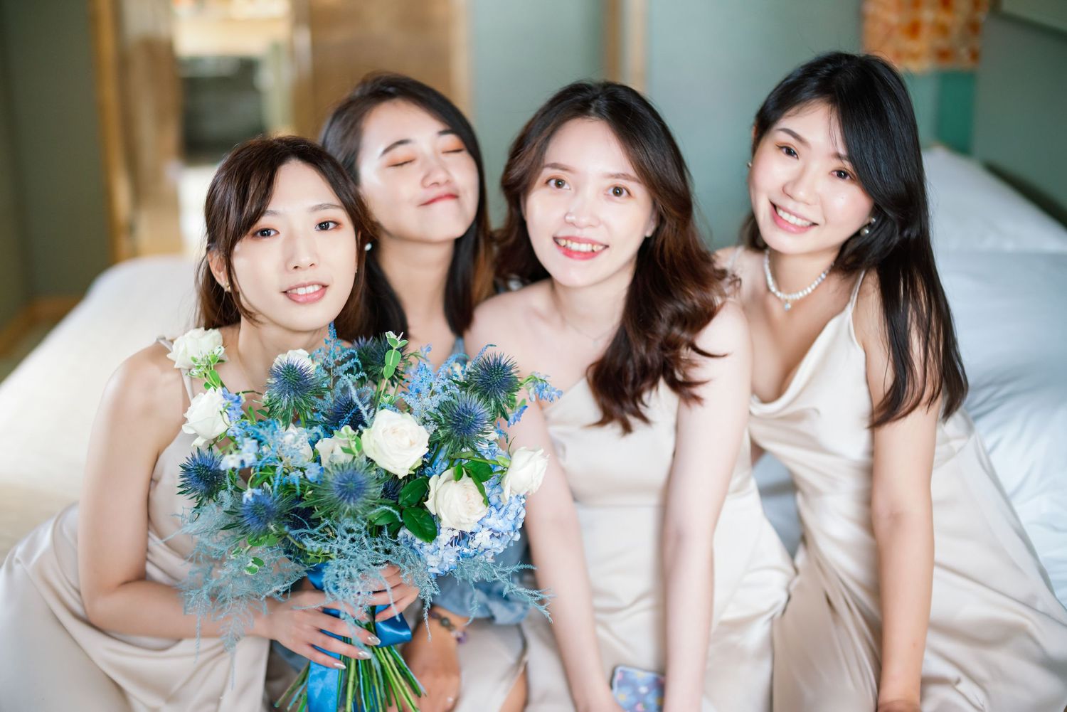 Group of women in light colored dresses posing together on bed with blue and white flower bouquet.