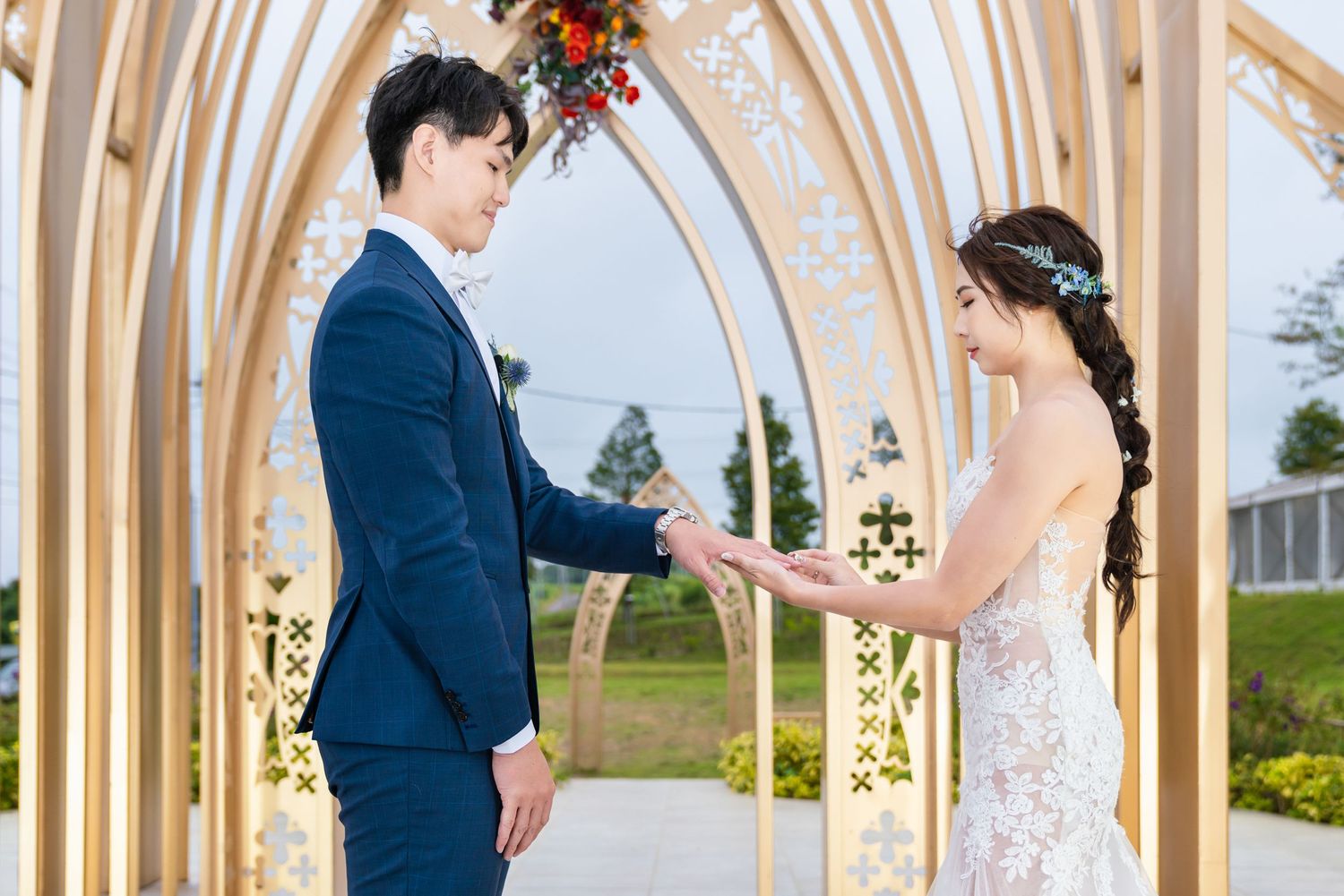 A couple exchanges rings during their wedding ceremony under an elegant arched pavilion decorated with flowers and lights.