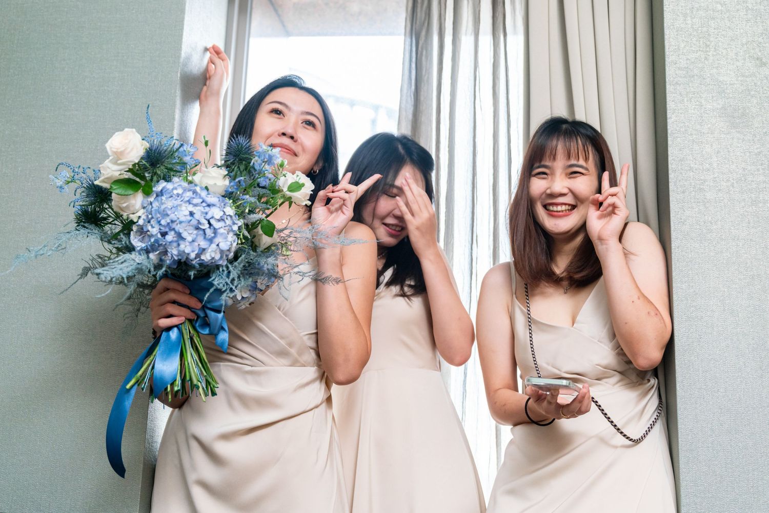 Three bridesmaids in light beige dresses celebrate joyfully with a blue hydrangea bouquet in a bright room.