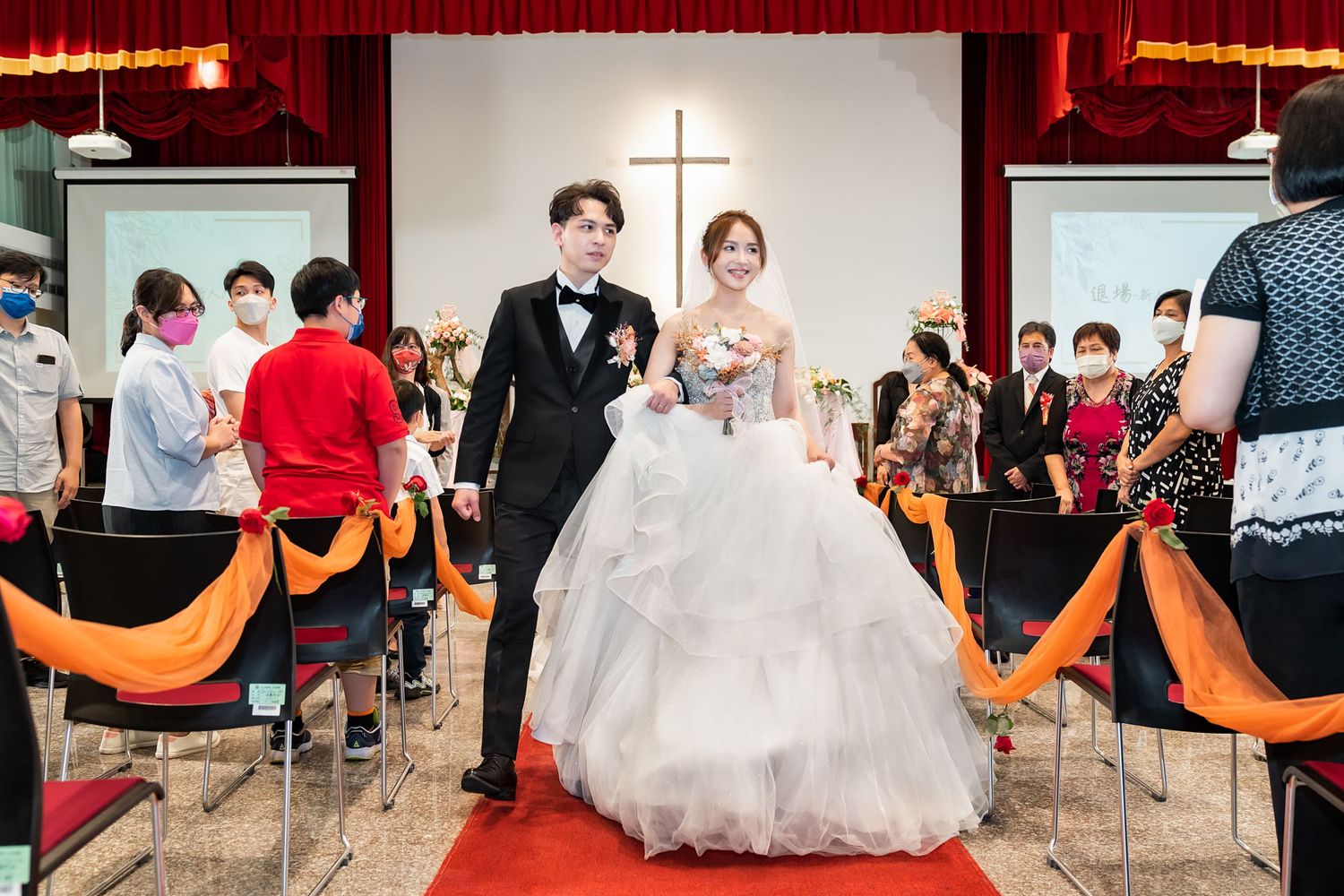 Wedding couple walks down church aisle decorated with orange fabric and a cross backdrop during ceremony.