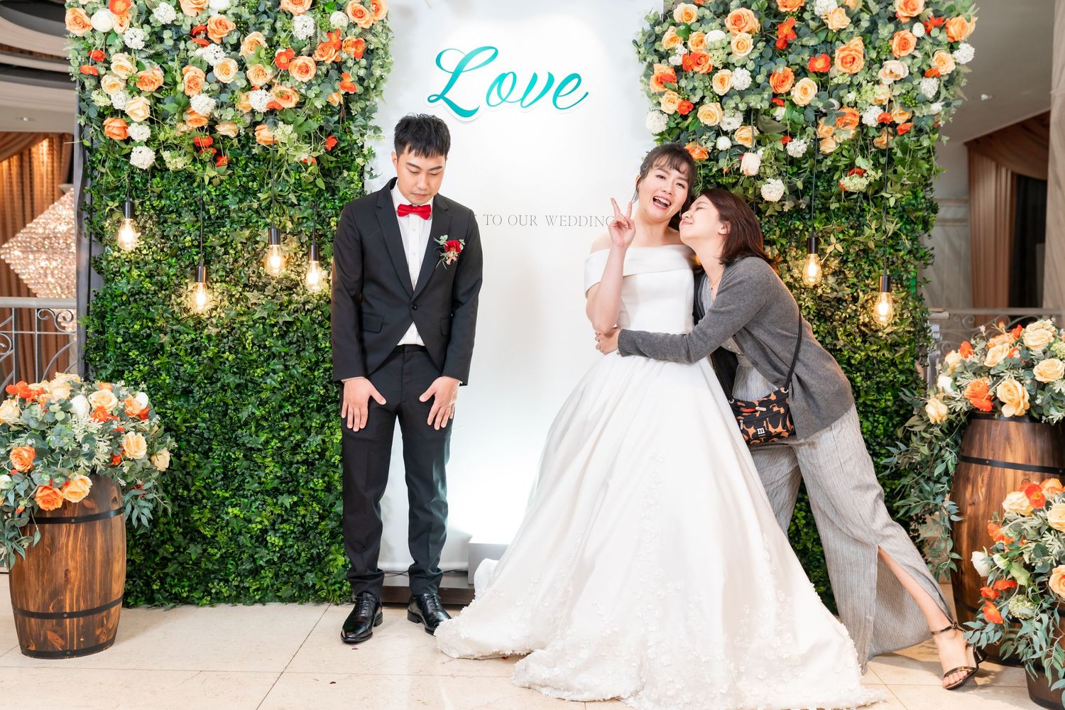 Playful wedding photo session with couple and guests in front of floral backdrop.
