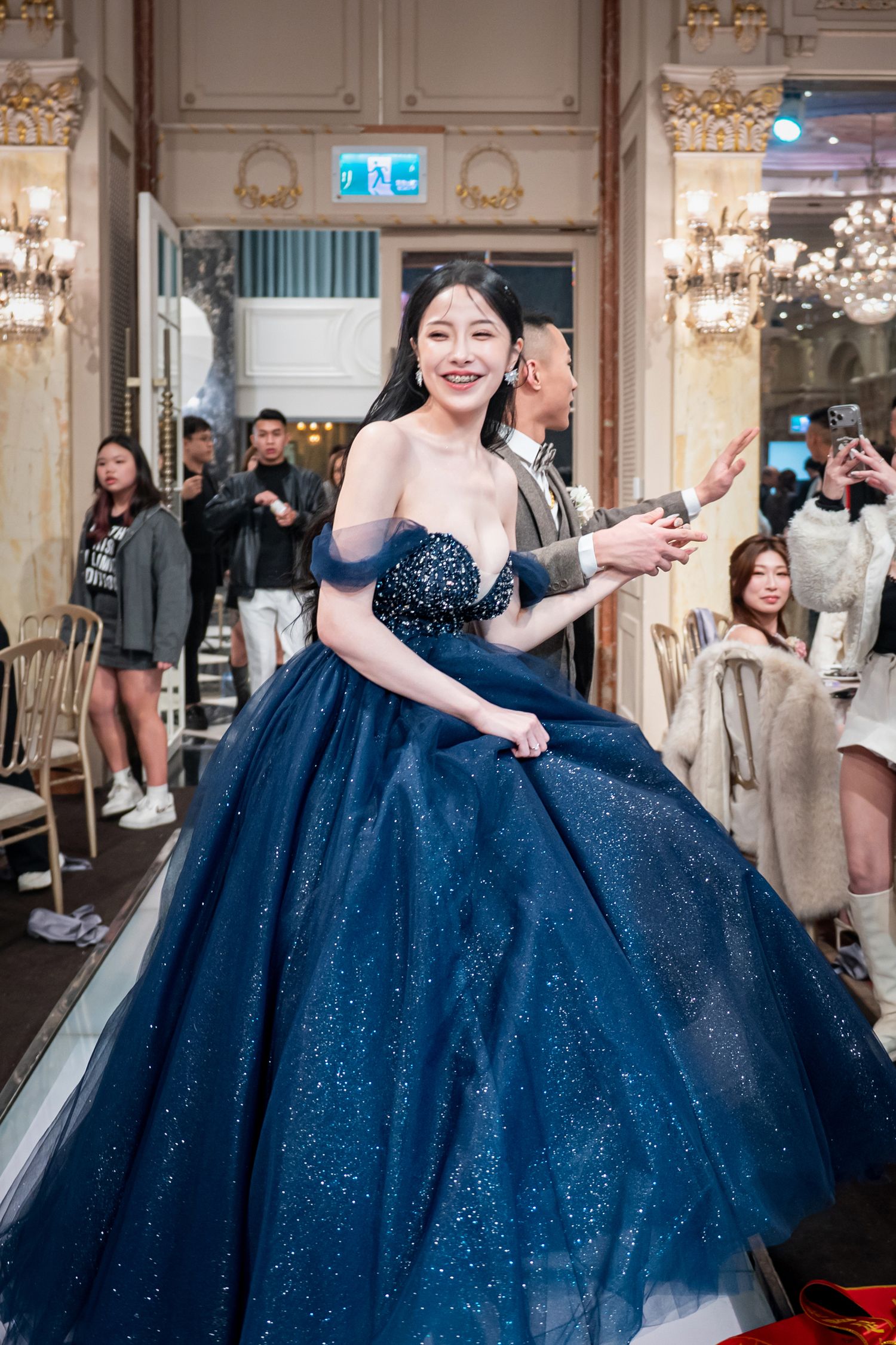 A person wearing a sparkling navy blue ball gown with off-shoulder design stands in an elegant venue with chandeliers.