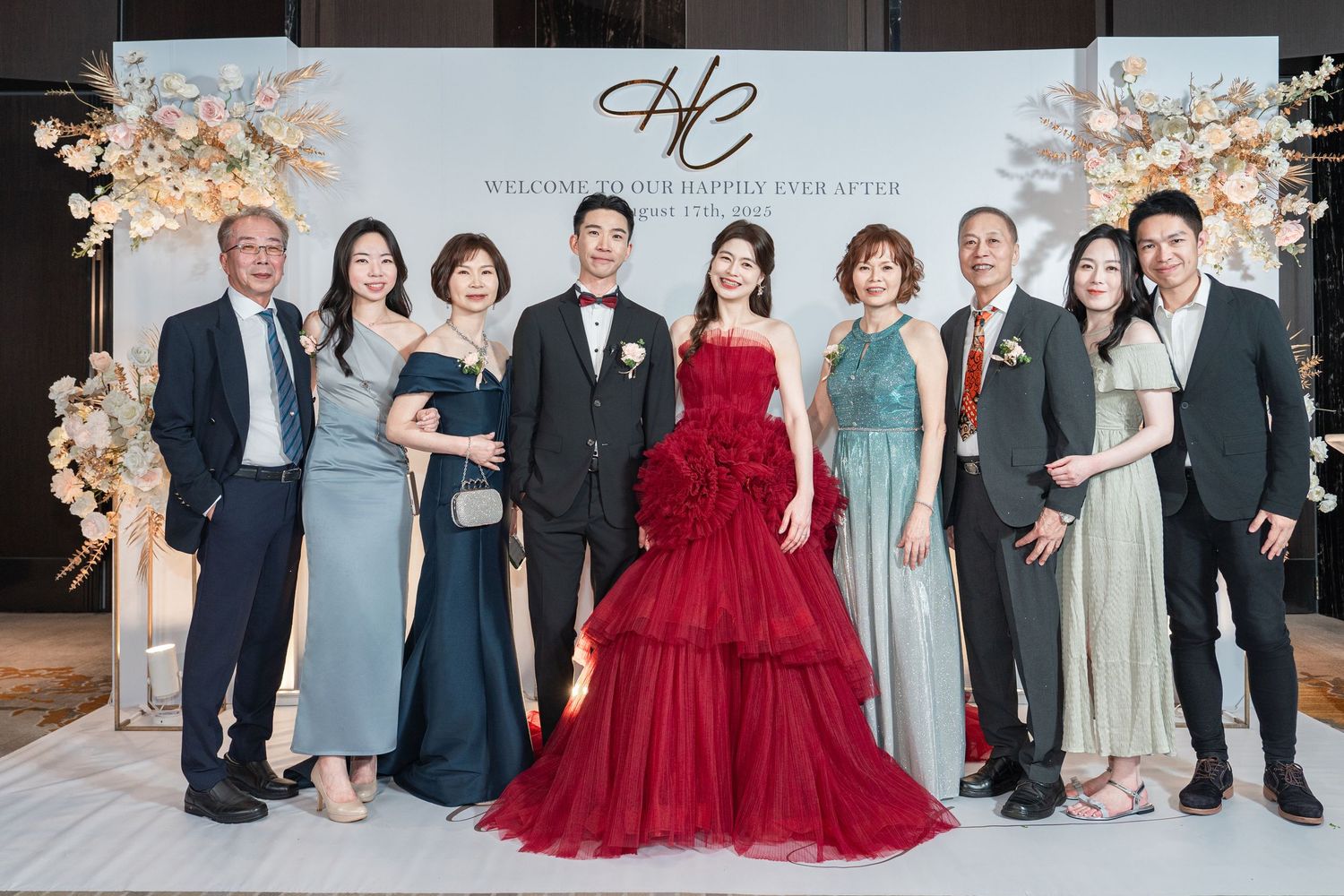 A wedding party poses together in formal attire with a red gown centerpiece against an elegant backdrop.