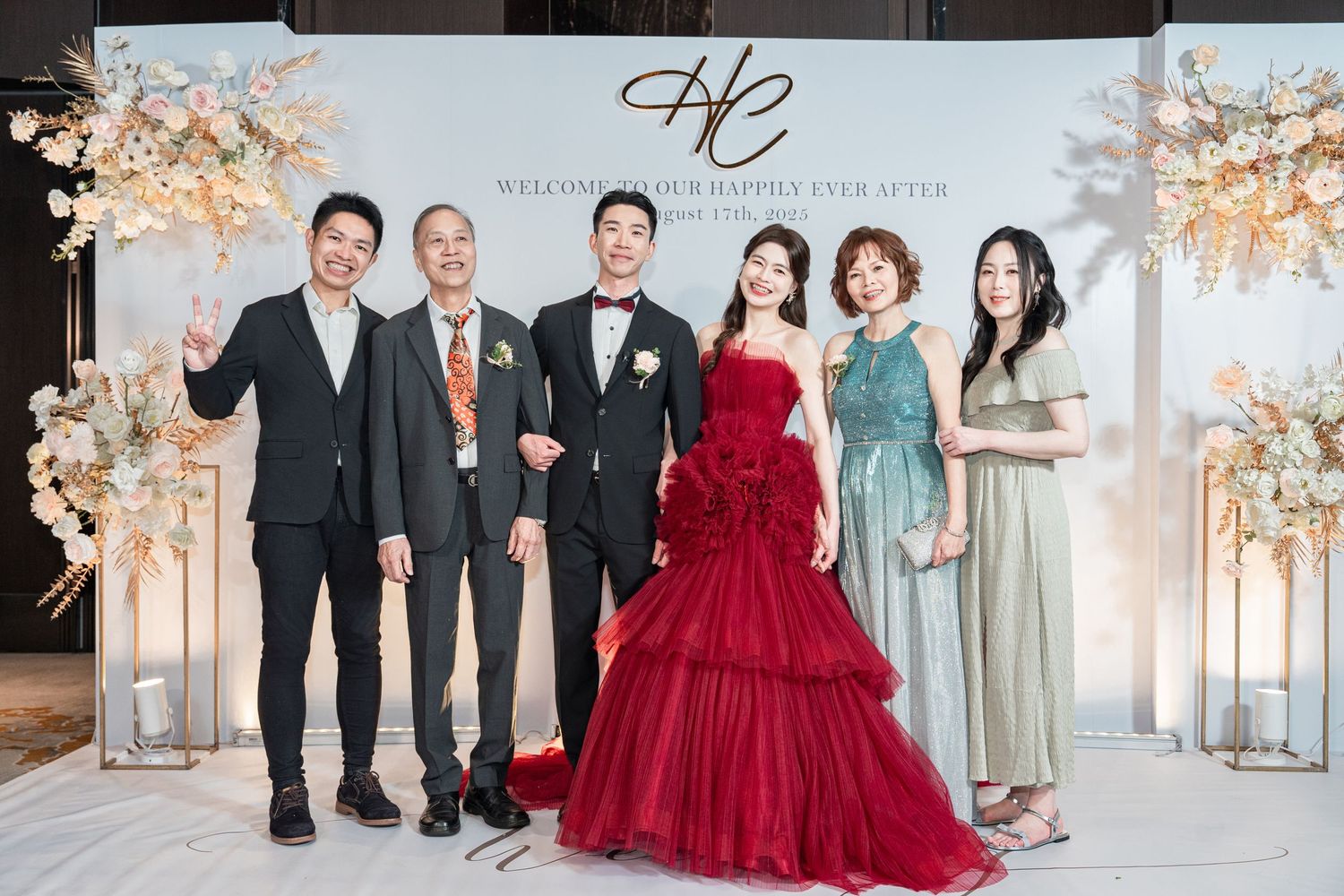 A group of formally dressed wedding guests pose in front of an elegant floral backdrop with white pillars and decor.