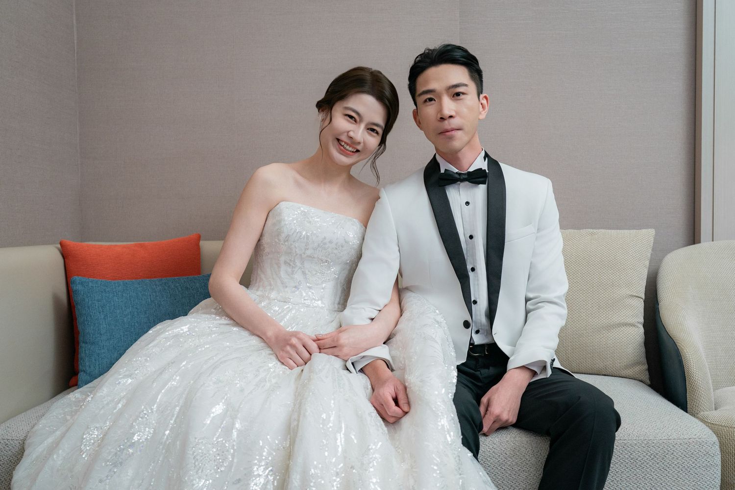 A couple in wedding attire sits together on a white couch in an elegant room setting.