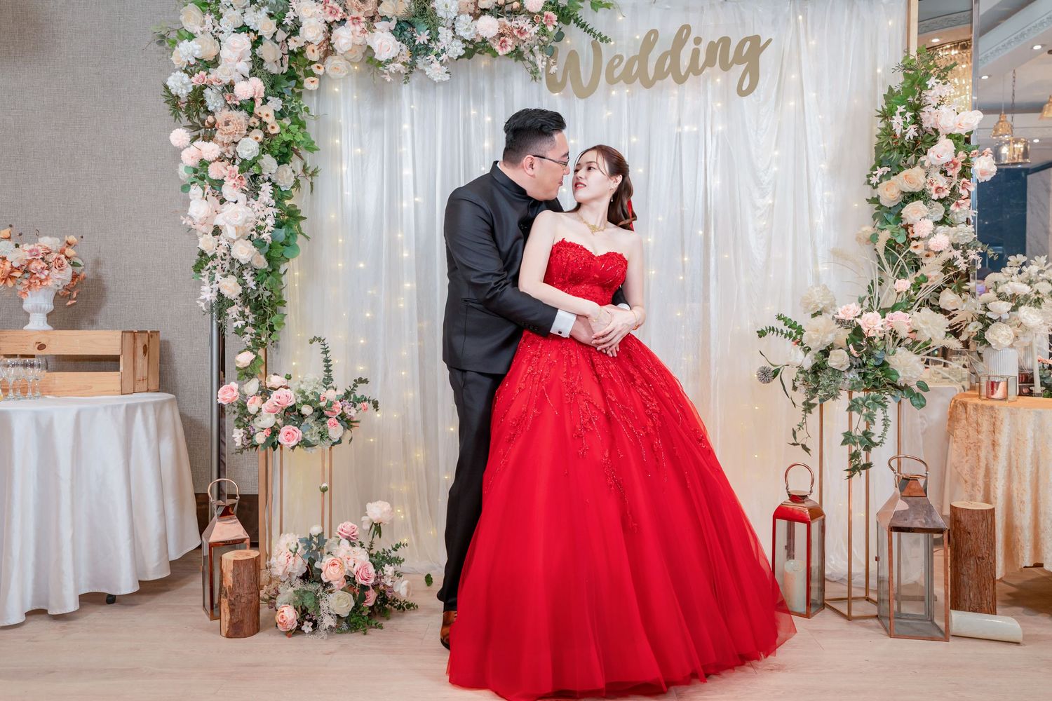 Elegant wedding scene with couple embracing by floral arch, bride wearing stunning red ball gown against white backdrop.