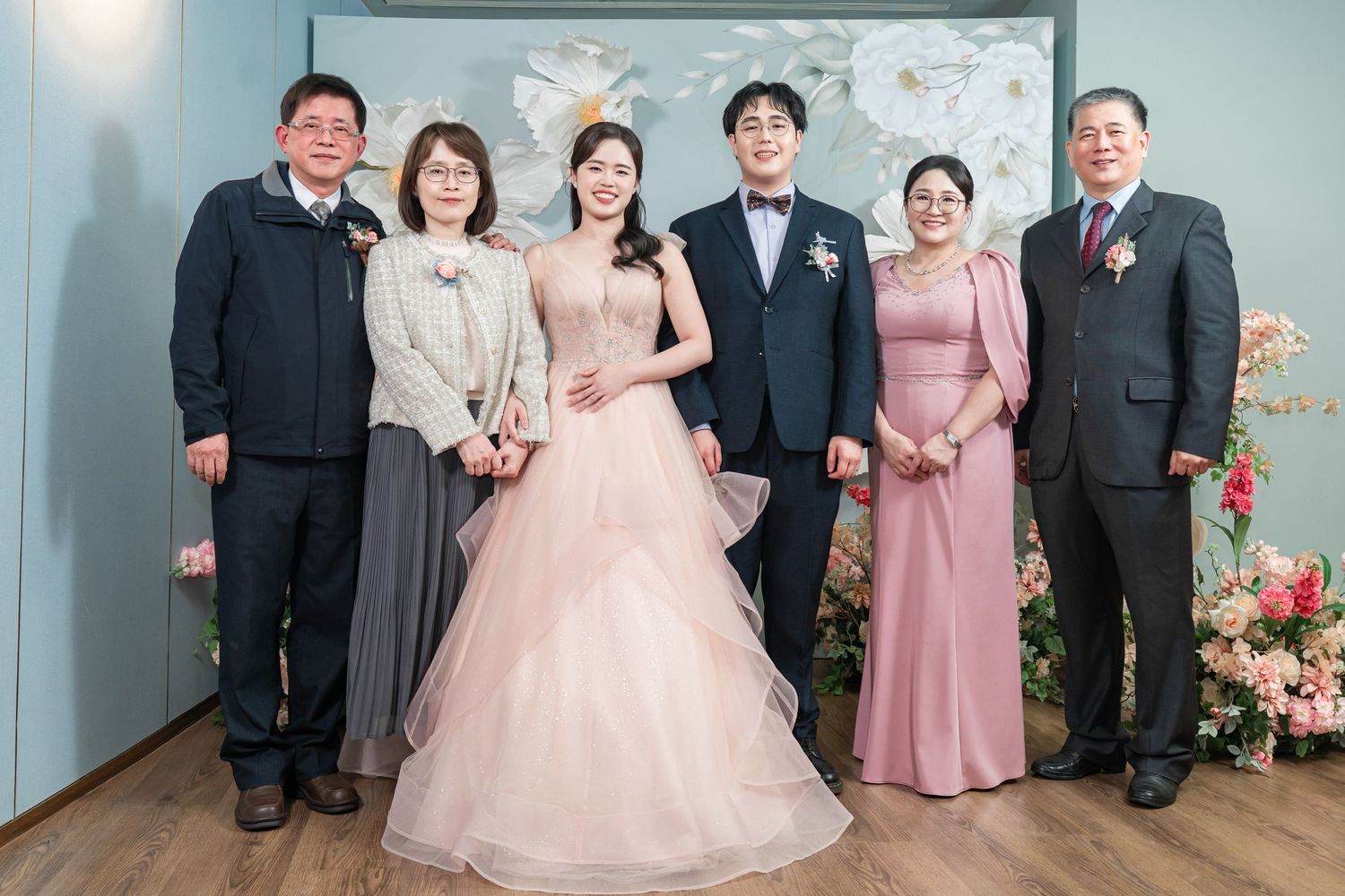 Wedding party in formal attire poses together against mint green wall with floral decorations.