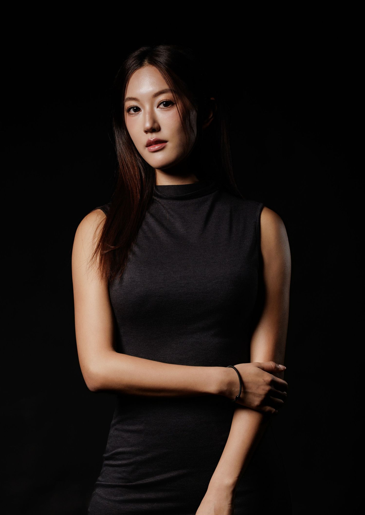 Young Asian woman in black sleeveless turtleneck poses confidently against dark studio background.